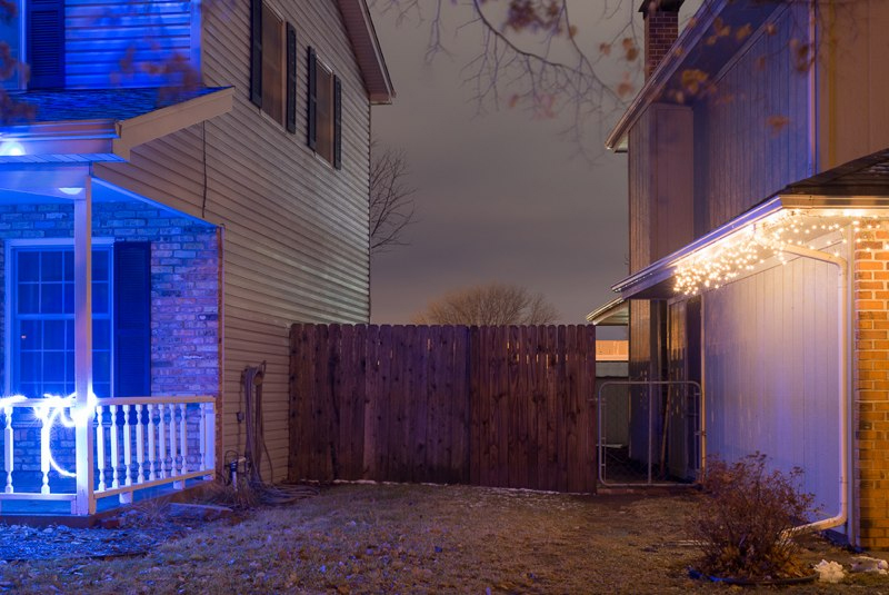 Nighttime photograph of residential houses with glowing neon blue and warm white lights illuminating a porch and garage, separated by a dark wooden fence in an otherwise dim suburban setting.