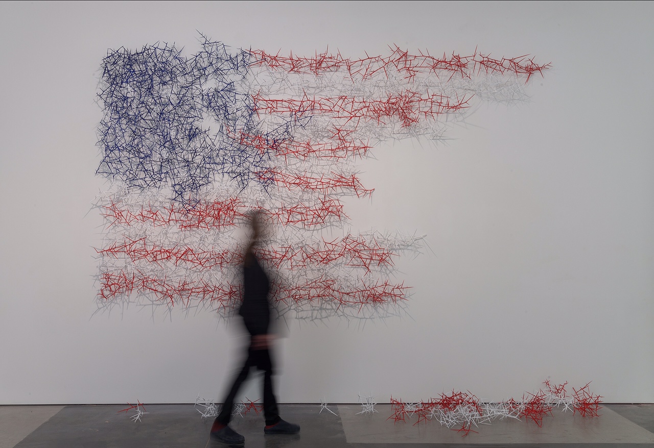 A blurred figure in dark clothing stands before a large abstract composition on a white wall made of interlocking spiky linear forms in navy blue, red, and gray. Scattered individual spike clusters rest on the floor below.