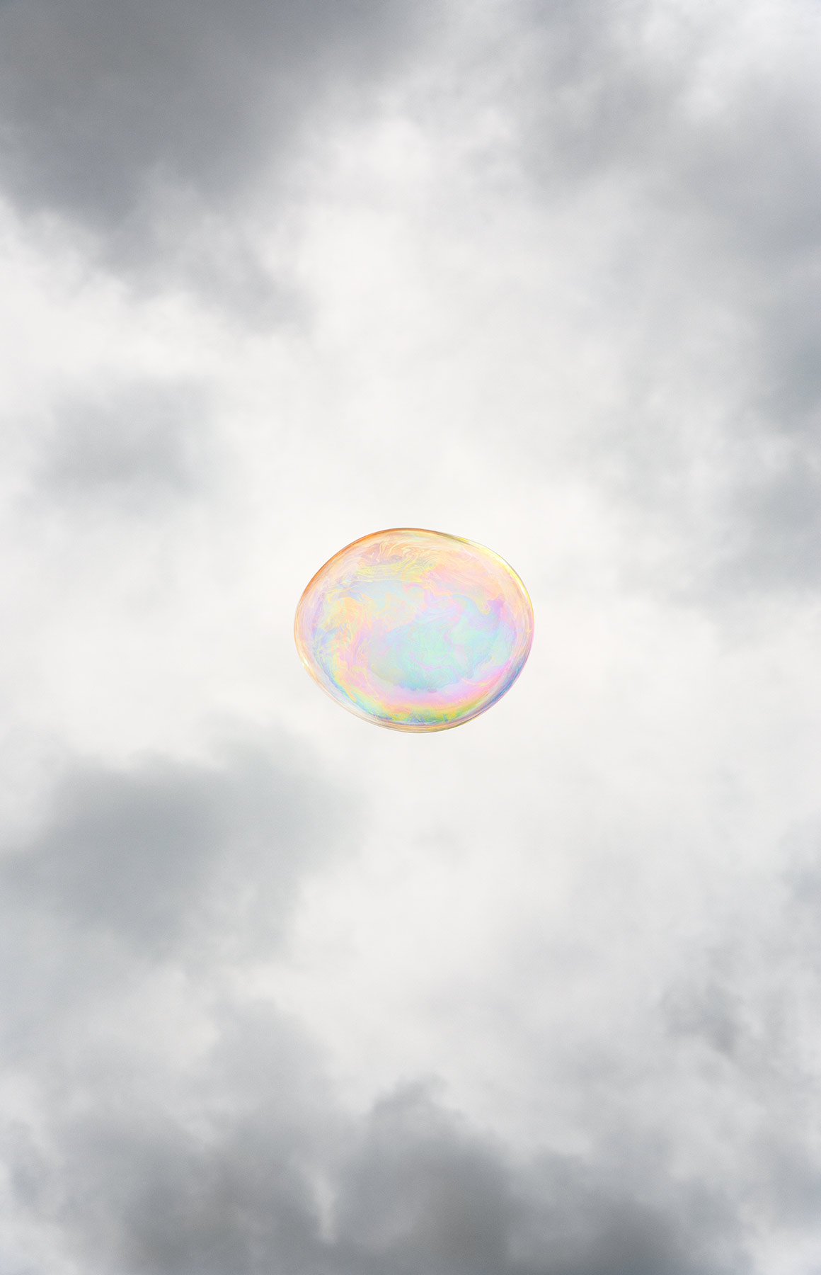A single iridescent soap bubble floats against a cloudy gray sky, its translucent surface displaying soft pastel hues of pink, yellow, and blue that shimmer across its spherical form.