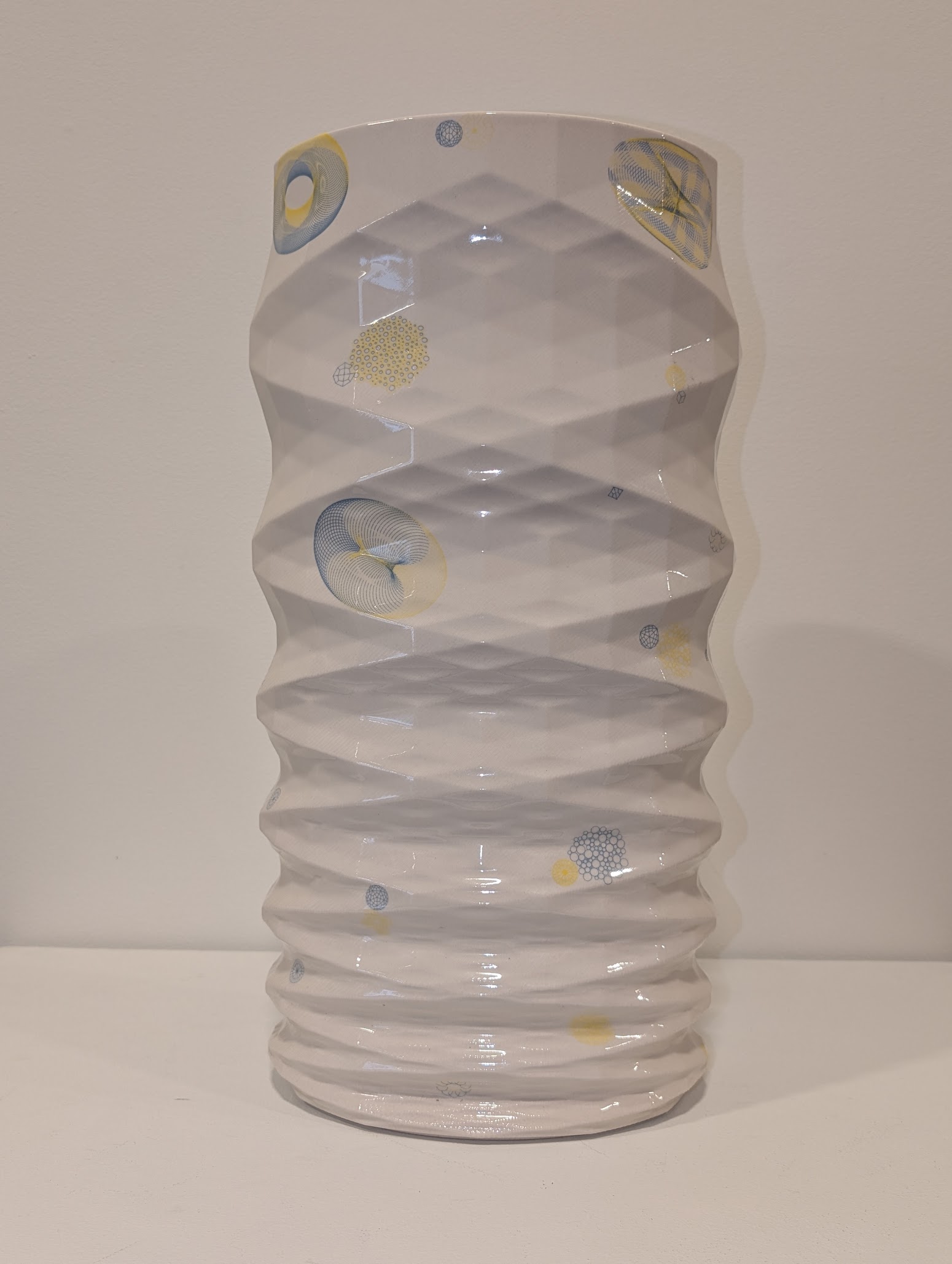 A white porcelain vessel with undulating, organic sides features abstract motifs in soft blue and pale yellow throughout—circular forms, linear patterns, and gestural marks that evoke scientific or natural imagery. The glazed surface catches light across its textured, rhythmically rippled form.