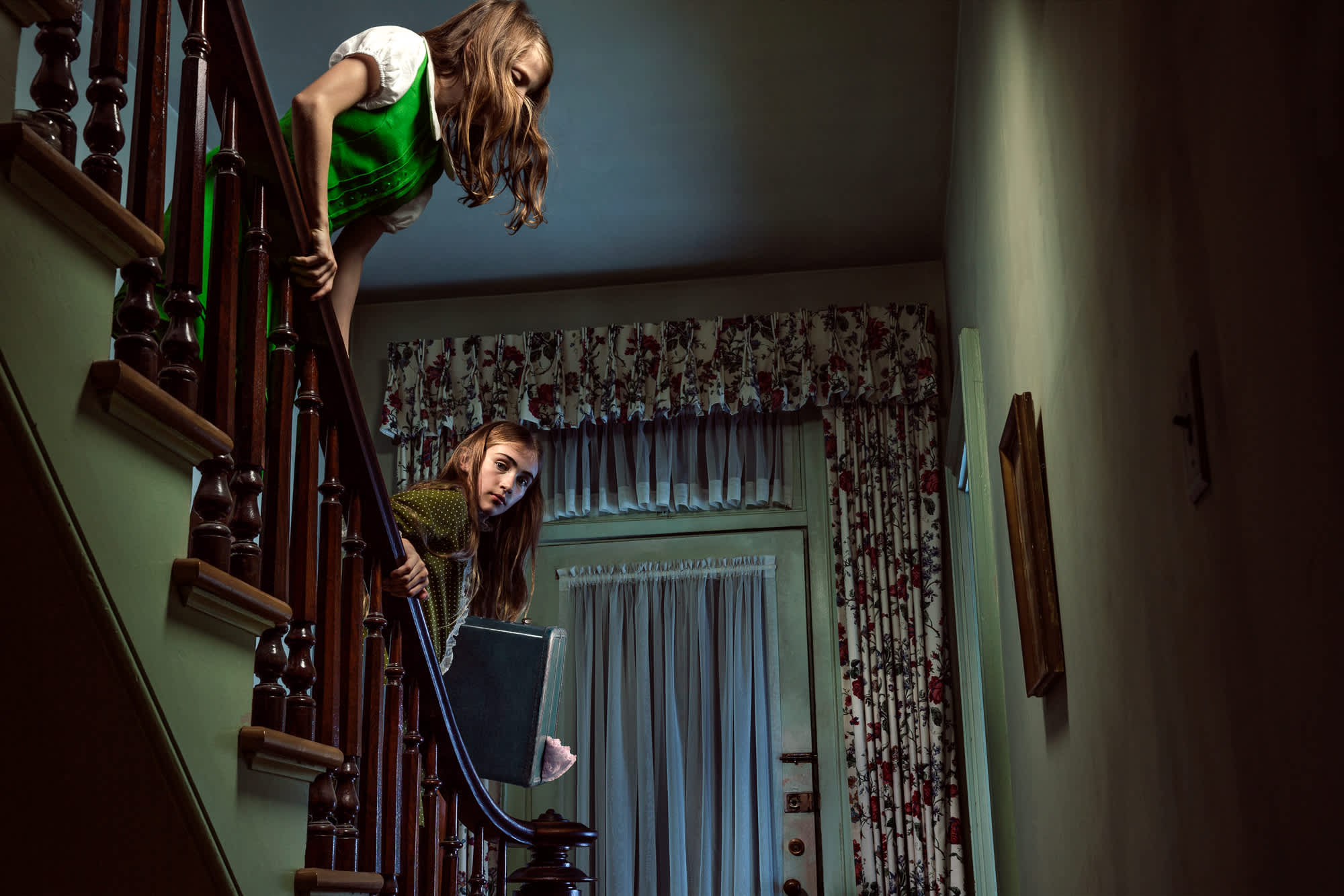 Two figures positioned on a wooden staircase in a mint-green interior with floral curtains and vintage furnishings. The upper figure wears a bright green garment and leans dramatically over the railing, while the lower figure peeks around the banister holding a dark object. Dramatic lighting and Dutch-angle perspective create a surreal, theatrical composition.