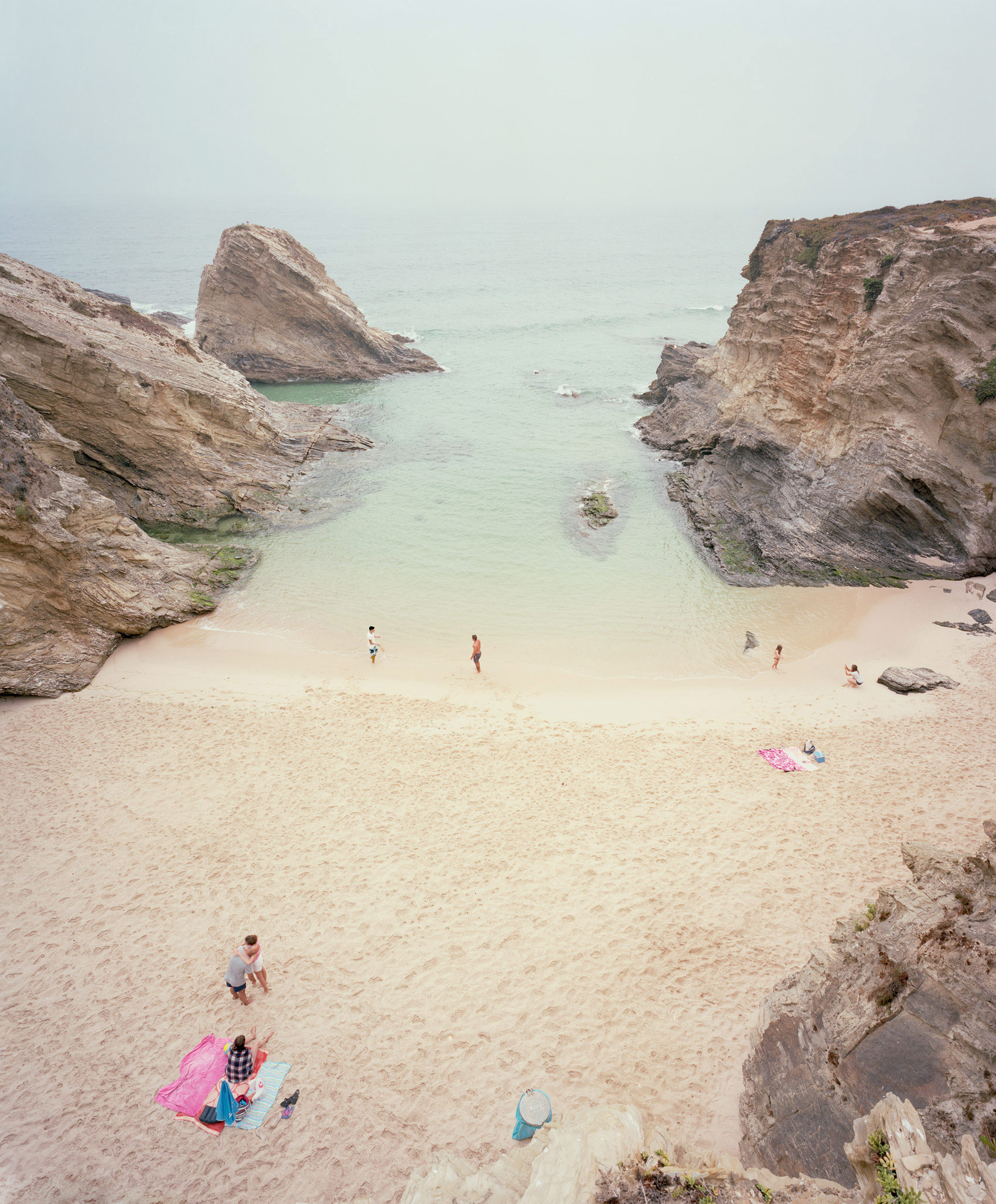 Aerial view of a secluded cove with pale turquoise water framed by dramatic, striated limestone cliffs. A small sandy beach is dotted with scattered beachgoers and colorful beach items, creating a serene summer scene rendered in soft, naturalistic tones.