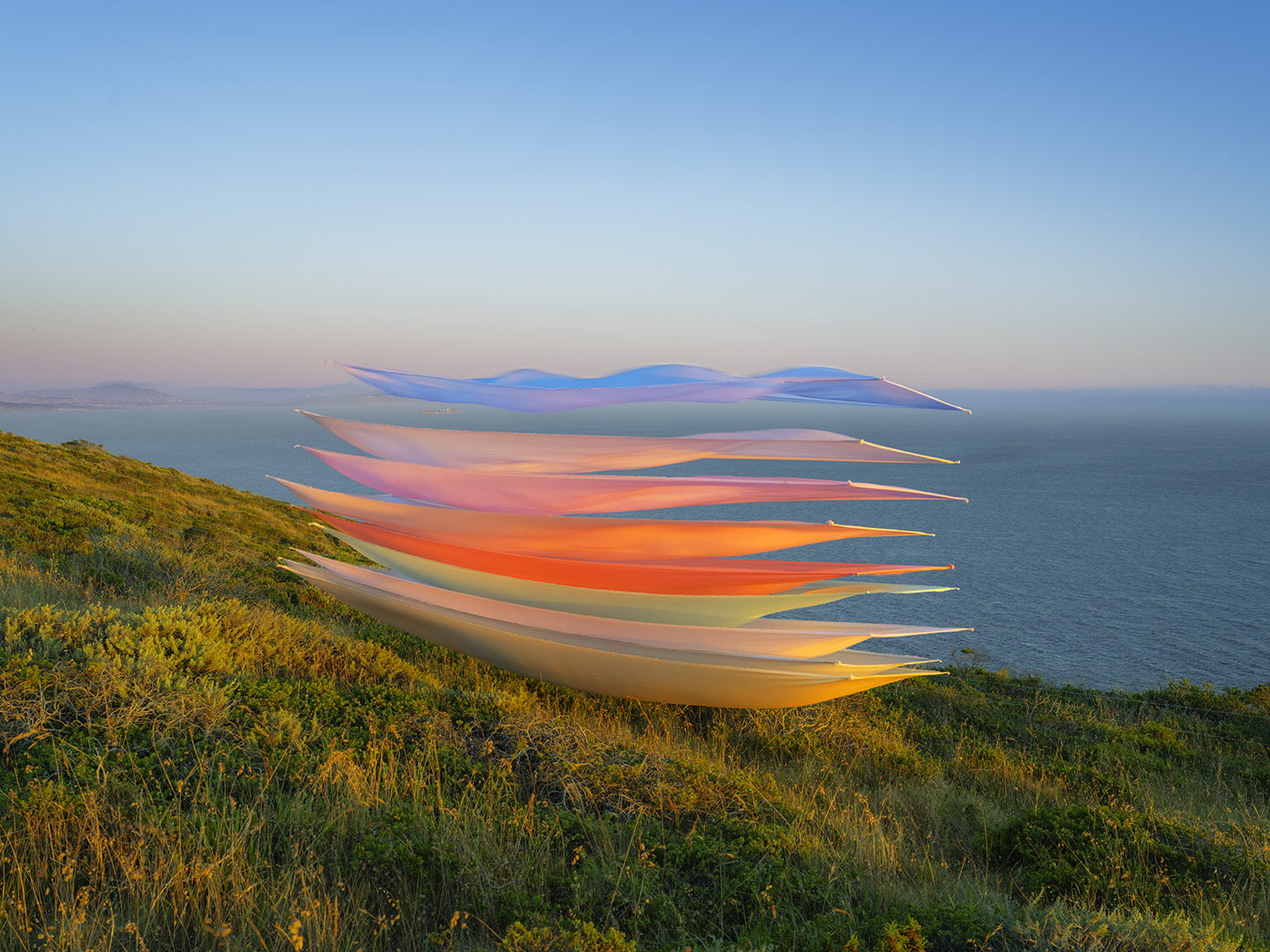 Layered silk ribbons in gradient hues—from deep blue at the top through pink, orange, and golden yellow—float horizontally above a coastal seascape, creating an ethereal interplay between the delicate fabrics and the ocean landscape below. The composition is anchored by wildflower-dotted coastal bluffs in the foreground, with distant hills visible across the water.