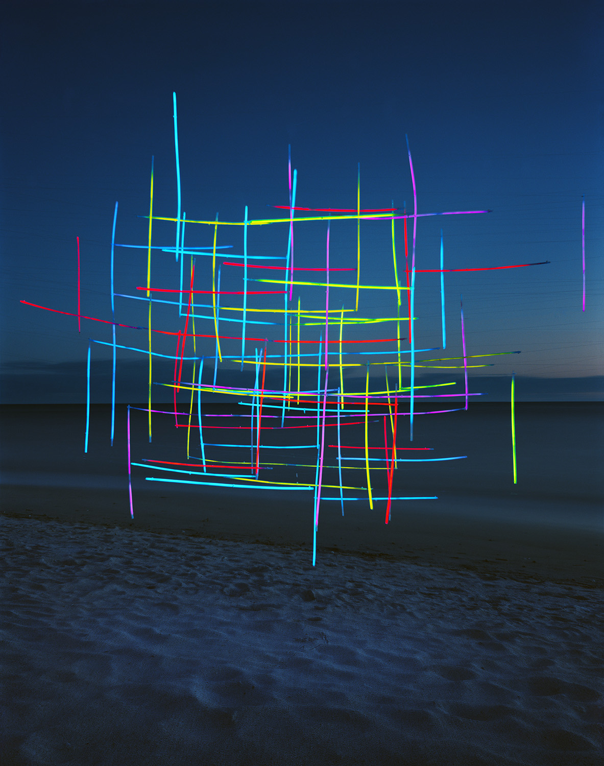 Neon-colored lines in cyan, yellow, red, magenta, and green form an intricate geometric lattice structure suspended on a beach at dusk, with the ocean and darkening sky visible behind it.