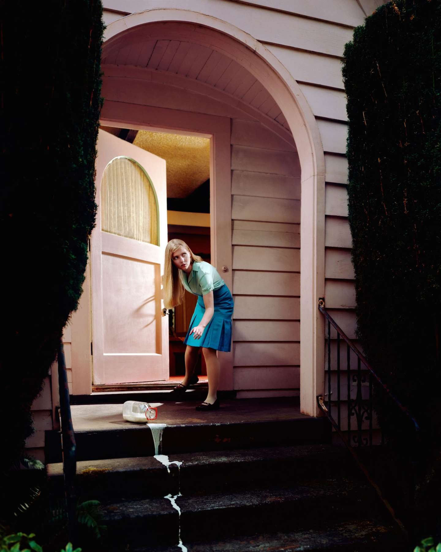 A woman in a blue dress and light green cardigan leans against a doorframe of a pale pink modernist house, gazing downward. Dark ivy-covered walls frame the arched entryway, while a spilled white liquid trails across the dark pavement in the foreground, creating a striking contrast against the warm, pastel architectural setting.