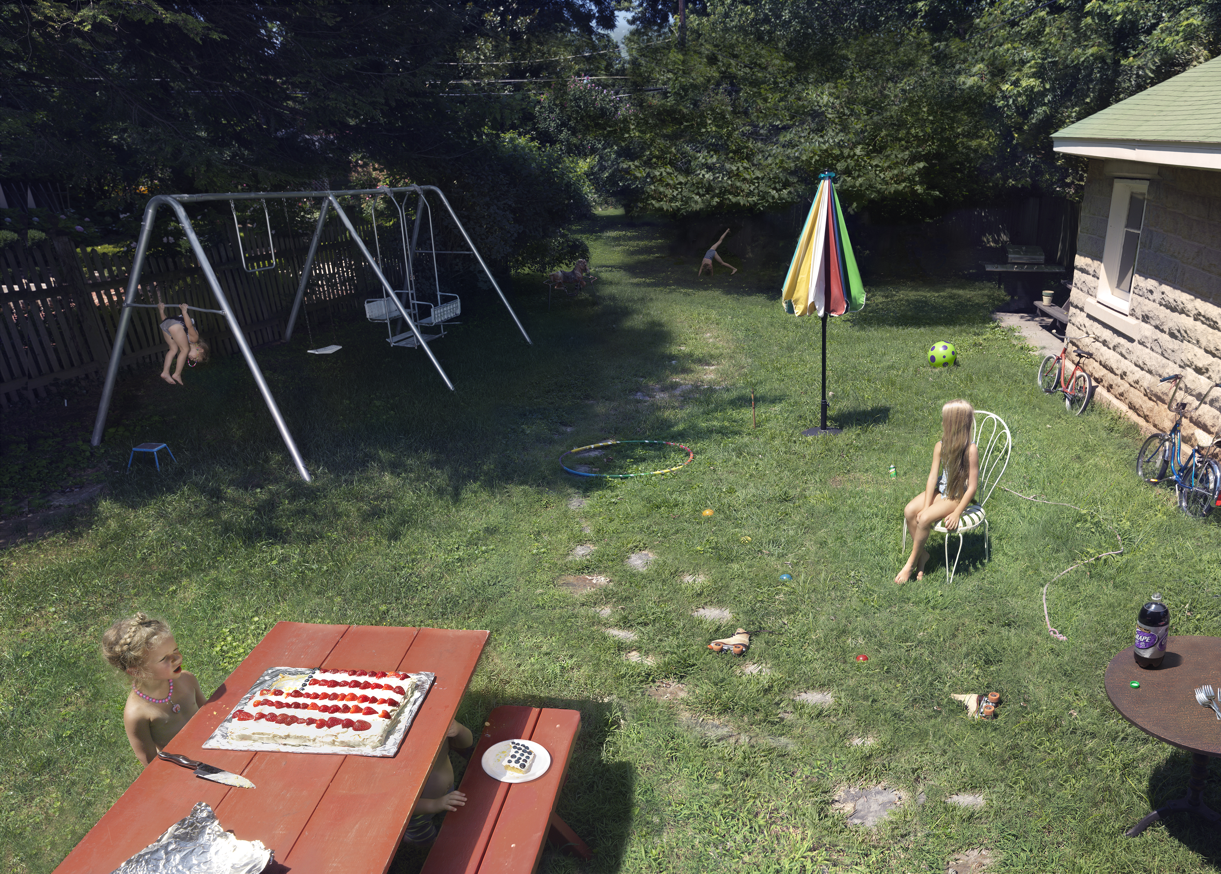 A backyard summer scene with children engaged in various activities: one child sits at a picnic table with a decorated cake, another sits on a chair in the middle distance, and a third swings on a metal swing set. Bicycles lean against a stone wall near a house, a colorful umbrella stands in the grass, and toys are scattered throughout the sunny lawn bordered by trees.