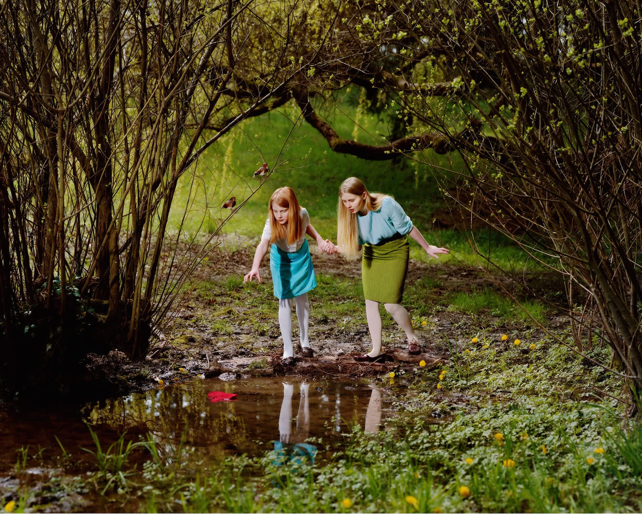 Two young girls with reddish-blonde hair stand on a narrow path beside a shallow stream in a dense woodland tunnel formed by overhanging branches. The girl on the left wears a teal dress with white socks, while the girl on the right wears a light blue shirt and olive-green skirt. Lush green foliage surrounds them, with small yellow flowers visible along the banks, and the scene is rendered in rich, saturated colors with warm and cool tones creating depth.