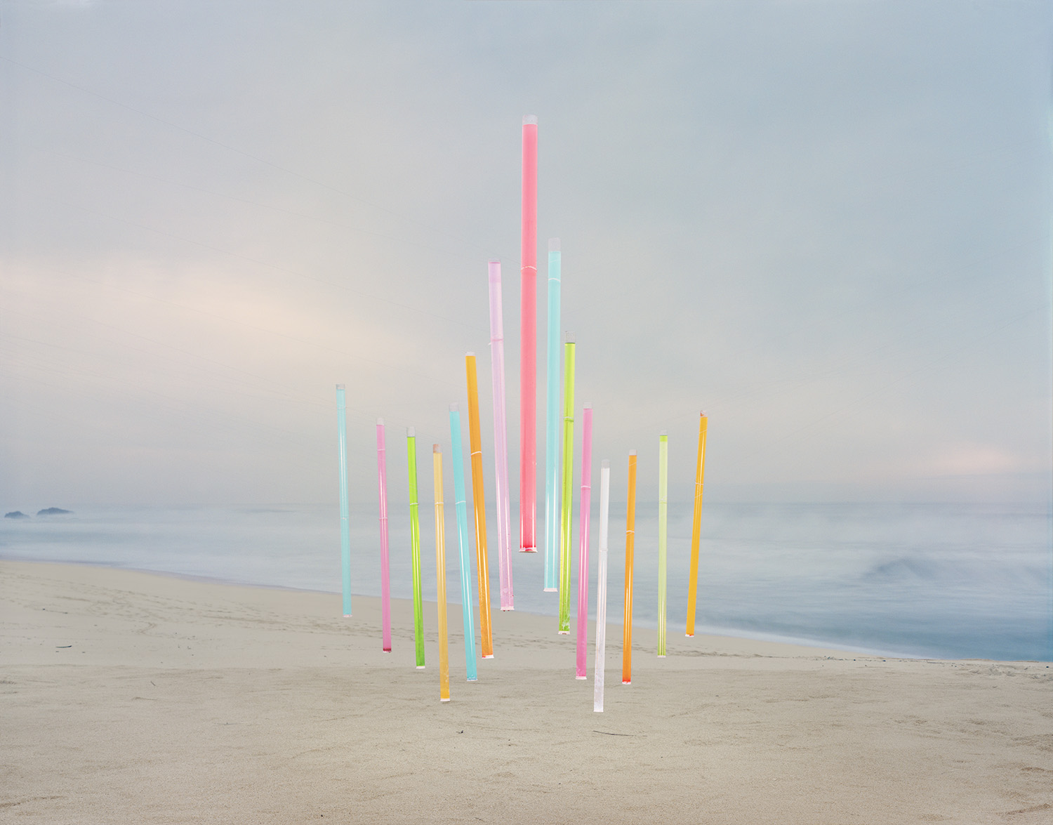 Vertical neon-colored tubes in pink, yellow, cyan, lime green, and white arranged in a clustered formation on a misty beach, with soft gray ocean and sky in the background.