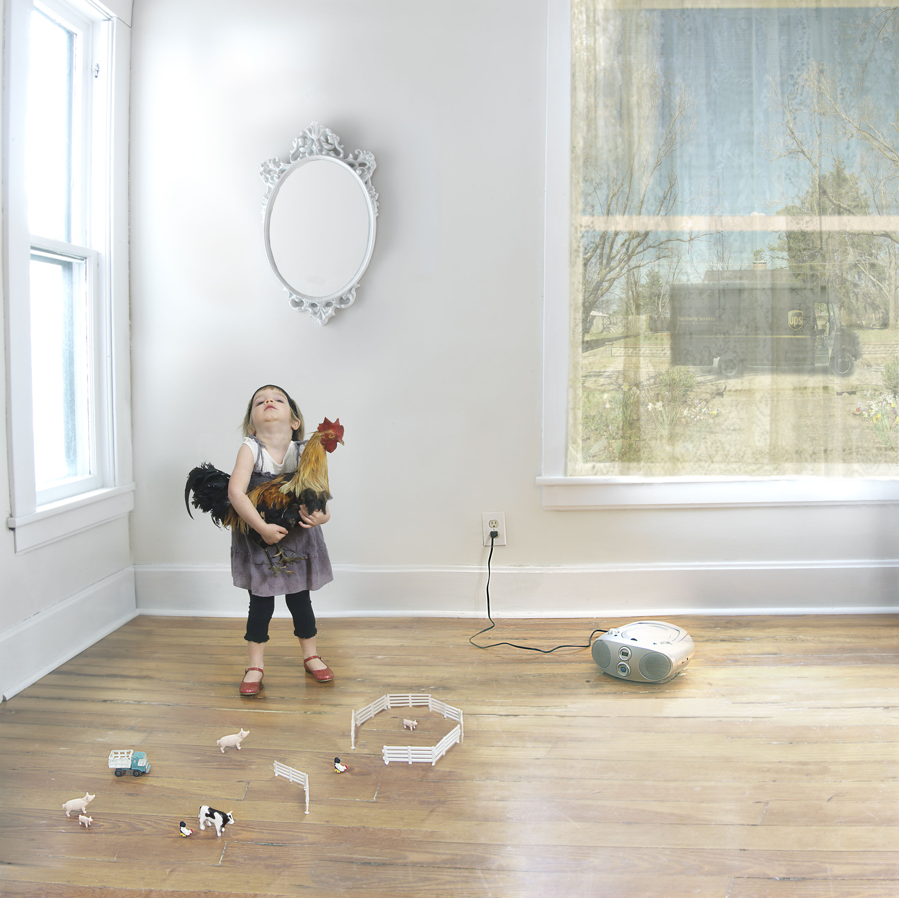 A young girl stands in a bright, minimalist room holding a rooster, gazing upward. Miniature toy farm animals and fencing are arranged on the wooden floor below her, alongside a portable speaker and electrical cord, creating a surreal domestic tableau bathed in natural light from large windows.