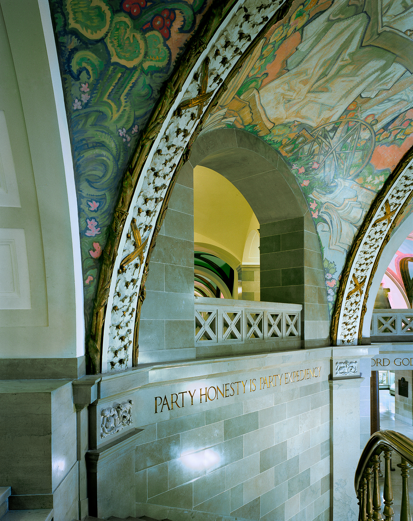 An ornate architectural passageway features a vaulted ceiling decorated with intricate green and gold scrollwork, floral motifs, and patterned borders. A stone wall below displays the text "PARTY HONESTY IS PARTY EXPEDIENCY" in gold lettering, while an arched doorway with a geometric lattice railing frames a view into an adjacent yellow-lit chamber.