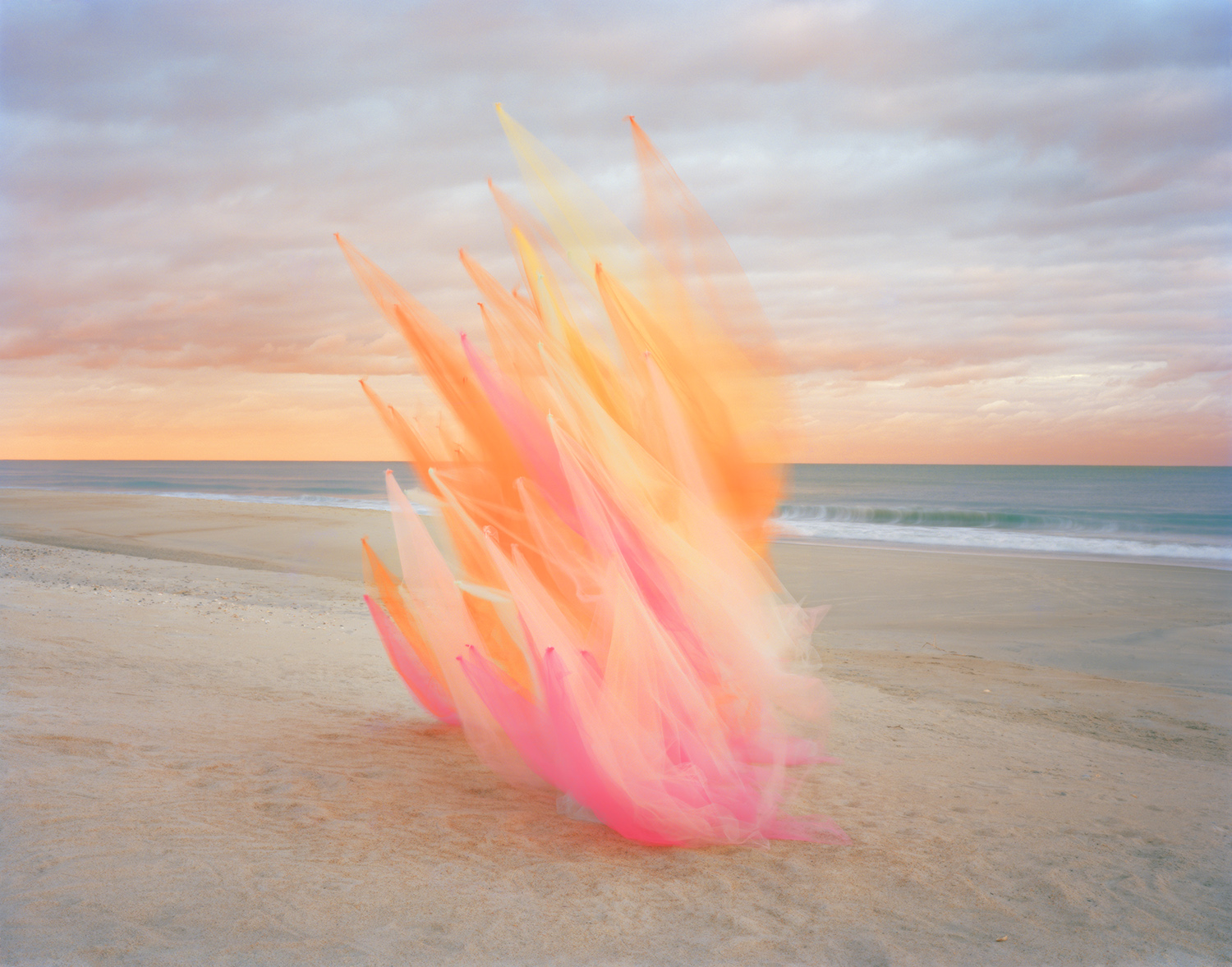 Translucent tulle fabric in shades of pink, orange, and yellow billows upward on a sandy beach at sunrise, creating an ethereal sculptural form against calm ocean waters and a soft, clouded sky.