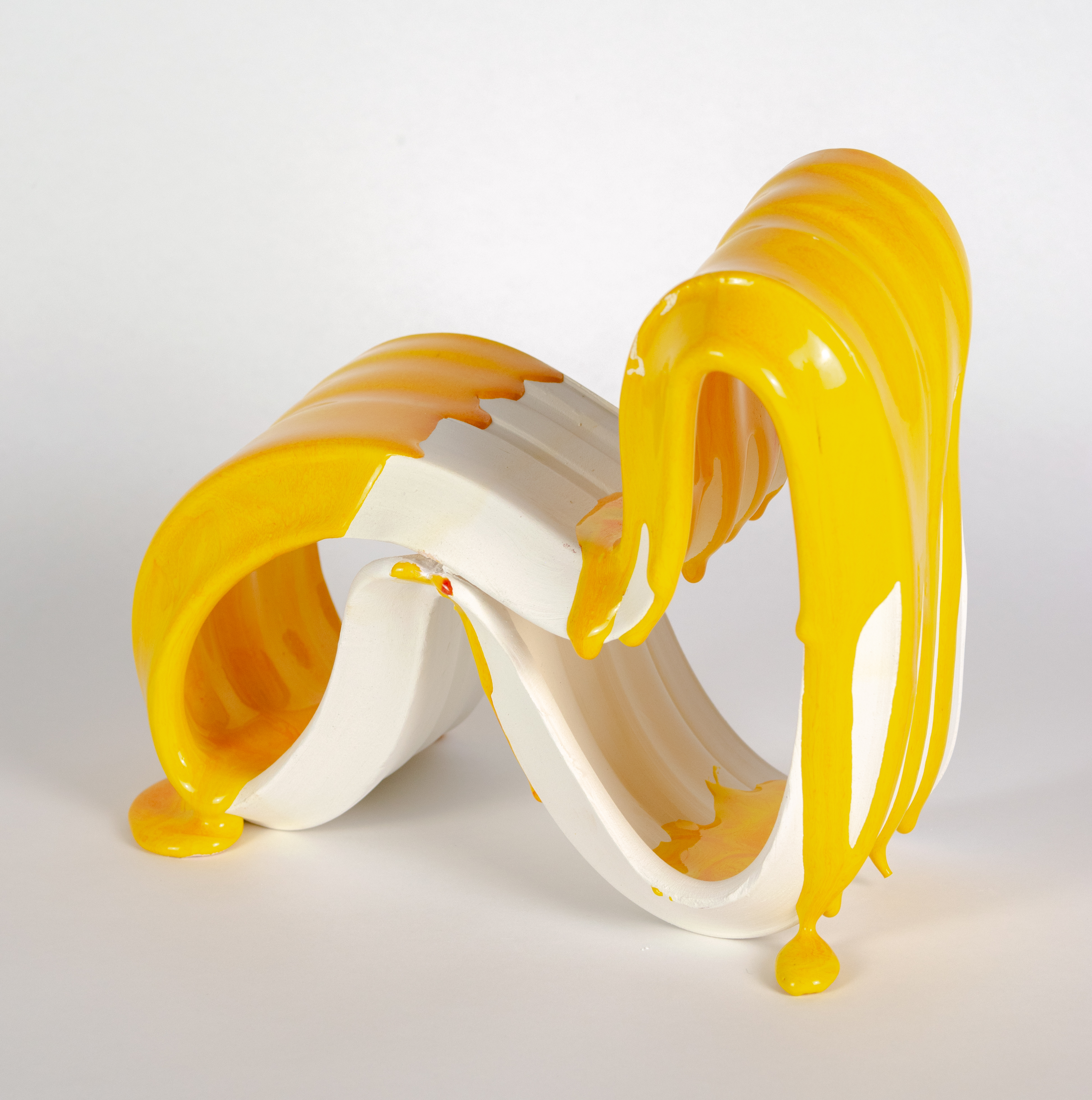 Abstract ceramic sculpture featuring curved white forms with glossy yellow elements flowing and draping across the composition. The piece sits on a neutral surface against a white background, showcasing dramatic contrasts between the matte white body and bright yellow glazed surfaces.
