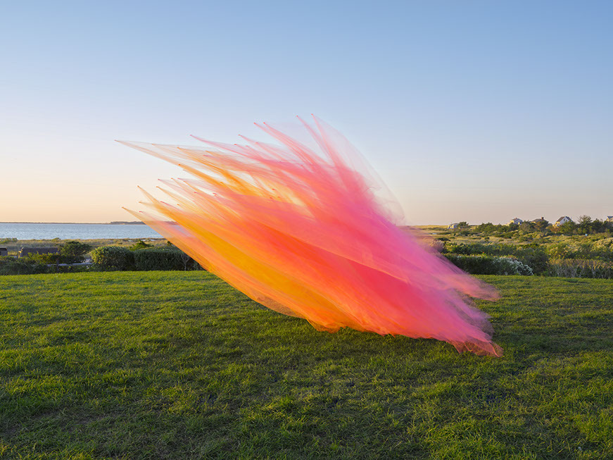 A sweeping diagonal plume of vibrant tulle fabric in gradient hues of orange, pink, and magenta billows across a green meadow overlooking a coastal landscape at dusk. The delicate, translucent material creates a dynamic sense of movement against the serene natural backdrop.