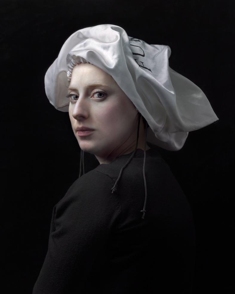 Portrait of a figure shown in three-quarter view against a black background, wearing a black garment with a distinctive white linen headwrap with voluminous folds. The figure gazes toward the viewer with a composed expression, rendered in a highly detailed, photorealistic style with naturalistic skin tones and subtle modeling of light and shadow.
