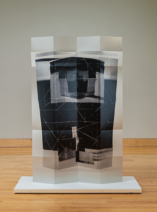 A translucent sculptural structure composed of layered panels arranged in a cubic form, featuring overlapping sheets of frosted and clear materials with geometric line drawings and dark photographic imagery visible through the layers. The work sits on a white base against a gallery wall with wooden flooring.