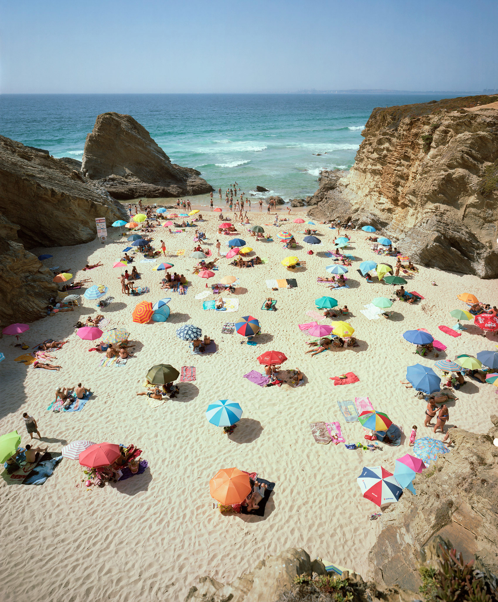 Aerial view of a crowded cove beach framed by dramatic rocky cliffs, with dozens of colorful umbrellas scattered across pale sand creating a vibrant patchwork pattern against the turquoise water and clear sky.