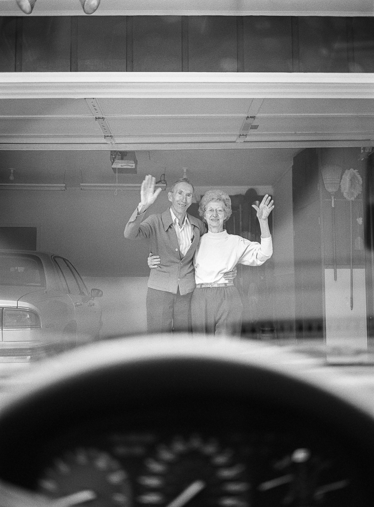 Black and white photograph taken from inside a car, showing a man and woman standing in a garage doorway waving goodbye. The couple is framed through the car's windshield with the hood visible in the foreground, creating a layered compositional depth.