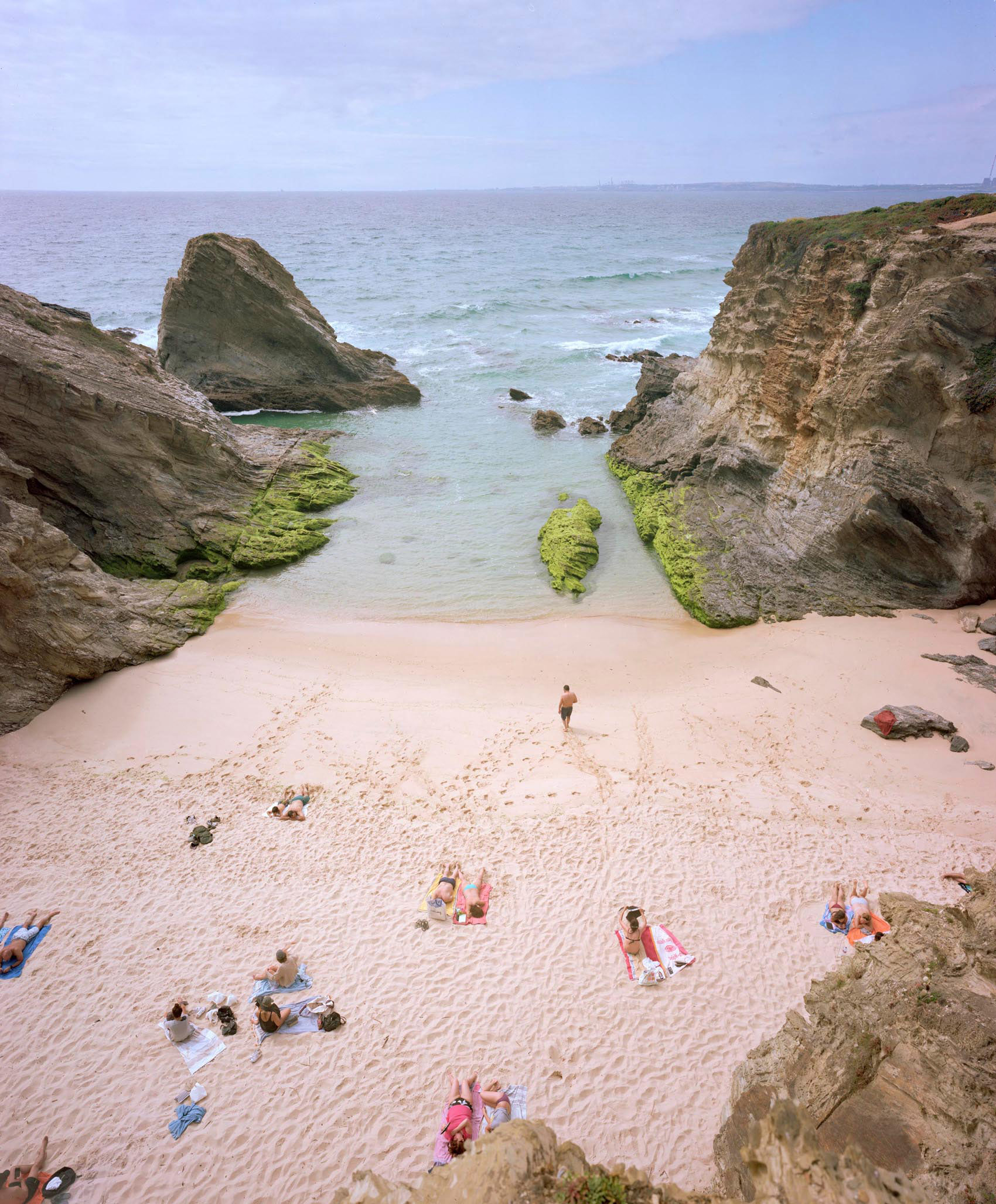 Aerial view of a secluded beach cove surrounded by dramatic moss-covered rock formations, with scattered beachgoers relaxing on pale sand and turquoise shallow waters dotted with seaweed-covered rocks.