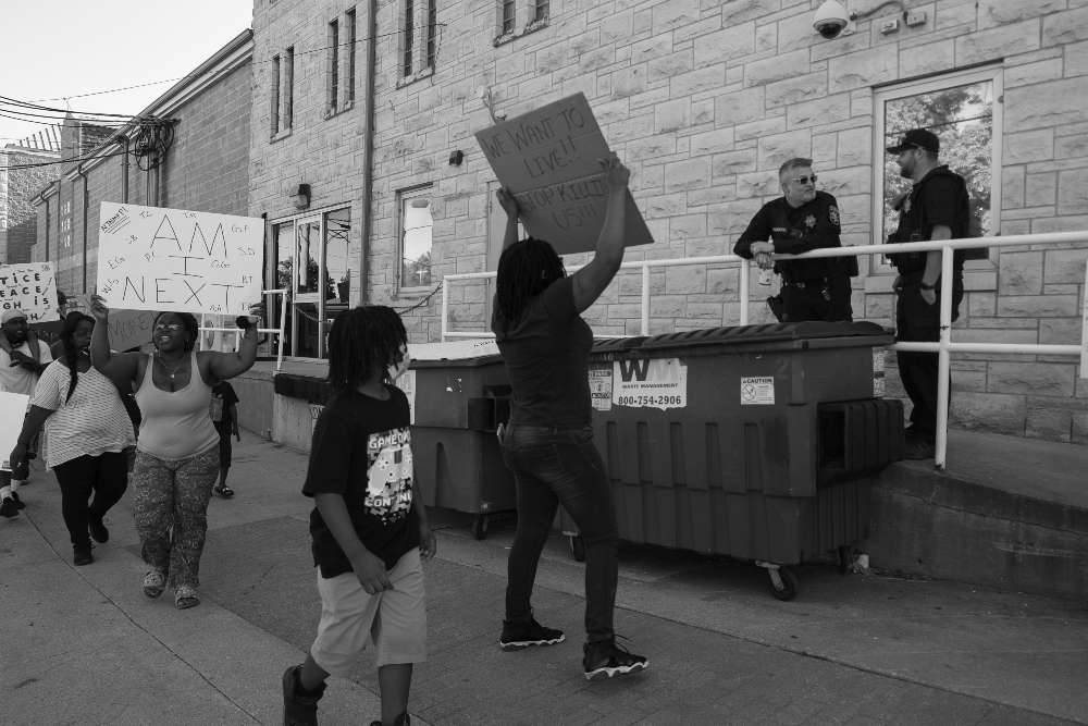 Black and white photograph of a street protest outside a brick building, showing demonstrators holding handmade signs while uniformed officers stand on an elevated platform. The composition captures the tension between the crowd at street level and the authority figures positioned above them on the structure.
