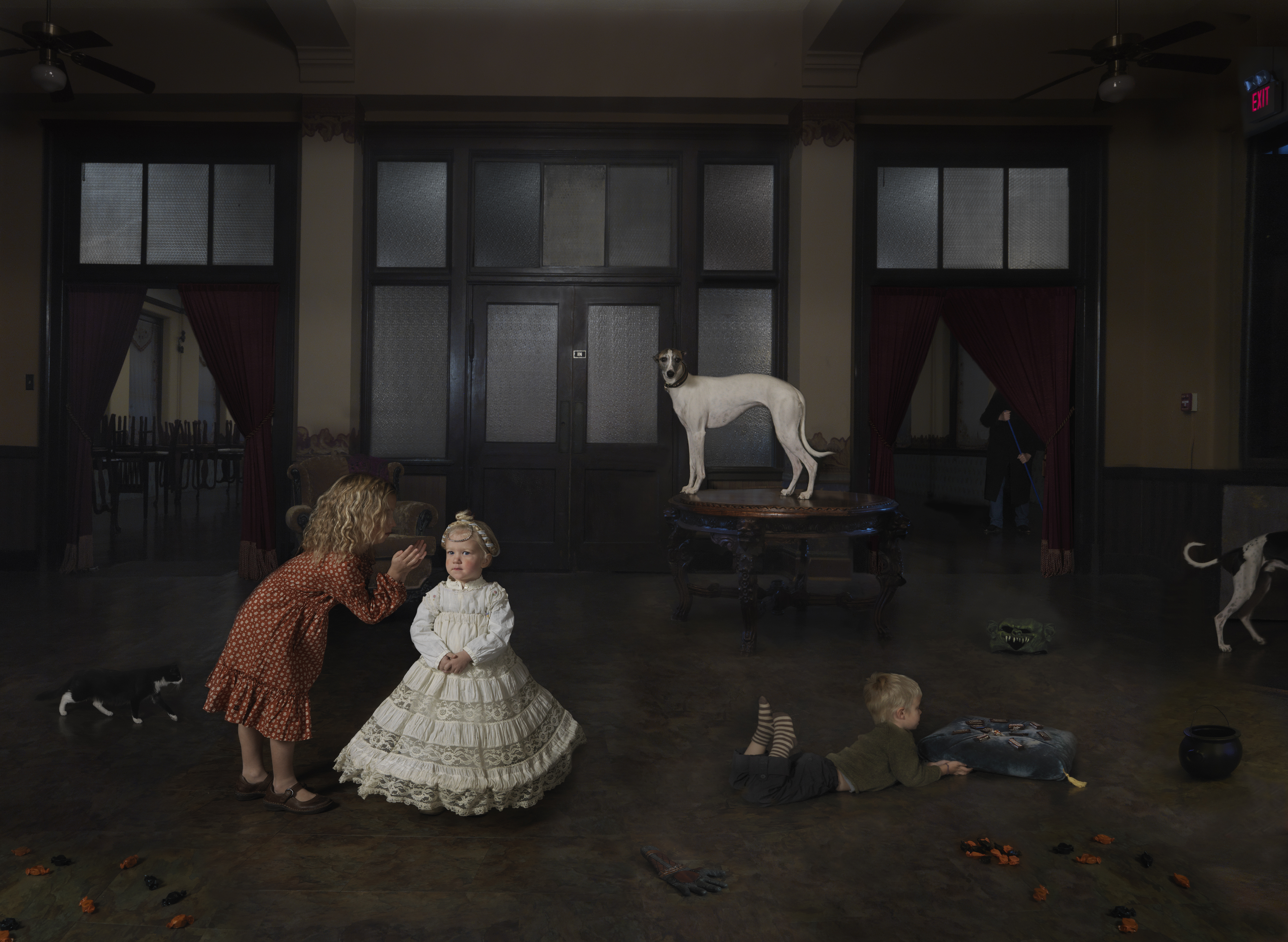 A surreal domestic scene depicts children and adults interacting in an industrial loft space with large windows and dark walls. A woman in a rust-colored dress bends toward a child in an elaborate white dress, while other figures and several dogs occupy the shadowy floor scattered with autumn leaves. A pale greyhound stands prominently on a platform in the center background.