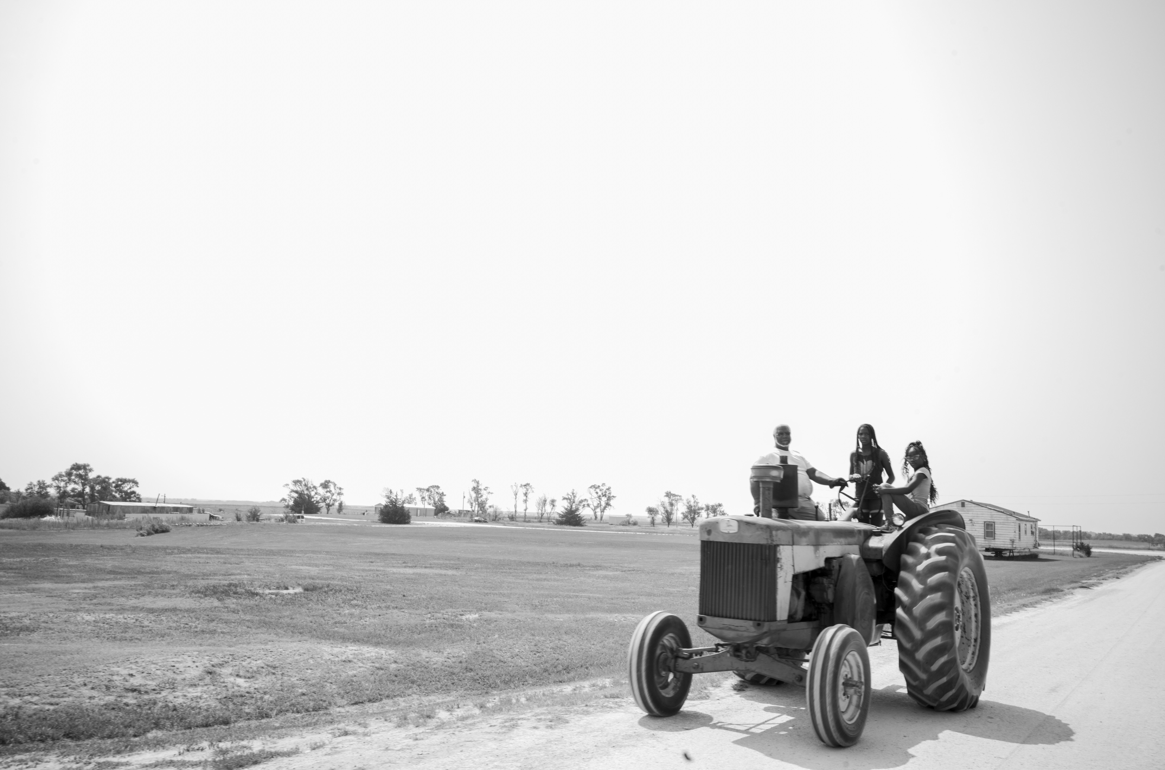Black and white photograph depicting three figures seated on a vintage tractor traversing a flat, rural landscape dotted with sparse trees and farm buildings along the horizon. The composition emphasizes the vast emptiness of the plains with the tractor positioned along a dirt road in the lower right portion of the frame.