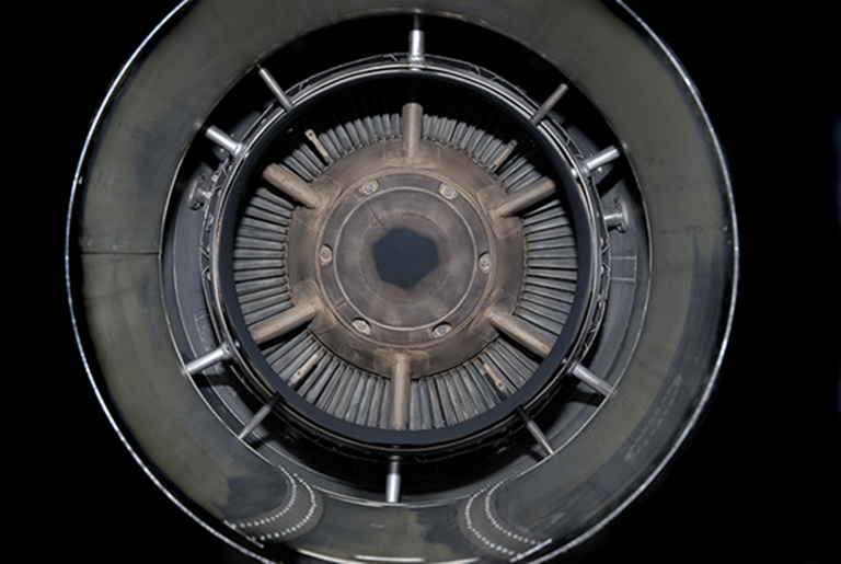 Concentric circular view of a mechanical turbine or engine intake, rendered in grayscale. Radiating fins and metallic components create a hypnotic, symmetrical pattern receding toward a dark central aperture, photographed directly from above against a black background.