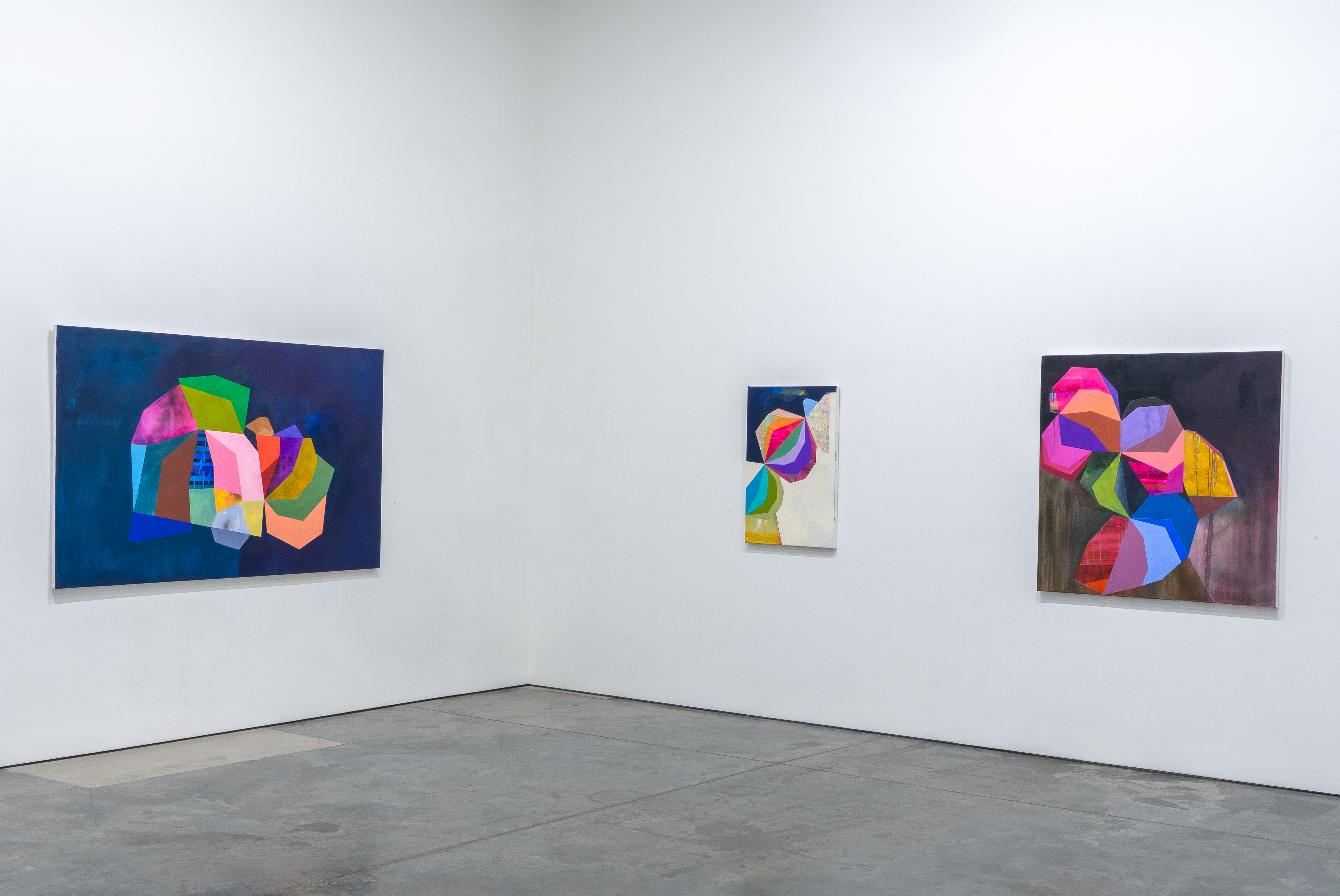 Gallery wall displaying three vibrant abstract artworks with bold geometric shapes in magenta, cyan, yellow, green, and orange against contrasting dark and light backgrounds, created through layered collage, acrylic, and oil techniques.