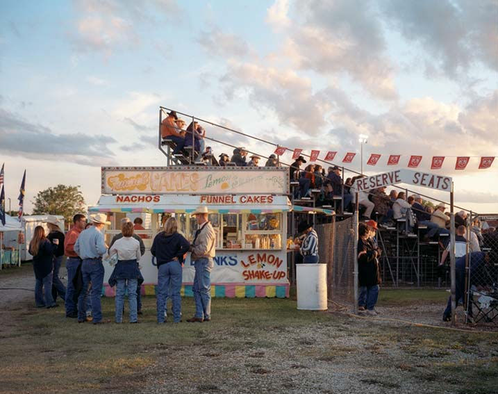 A fair or carnival scene at dusk showing a food vendor stand with signage for nachos, funnel cakes, lemon shake-up, and other refreshments. Visitors in casual clothing stand in front of the stand and queue on an elevated platform with "reserve seats" signage, beneath a string of red flags strung overhead. Grass grounds the foreground while a clouded sky occupies the upper portion of the composition.
