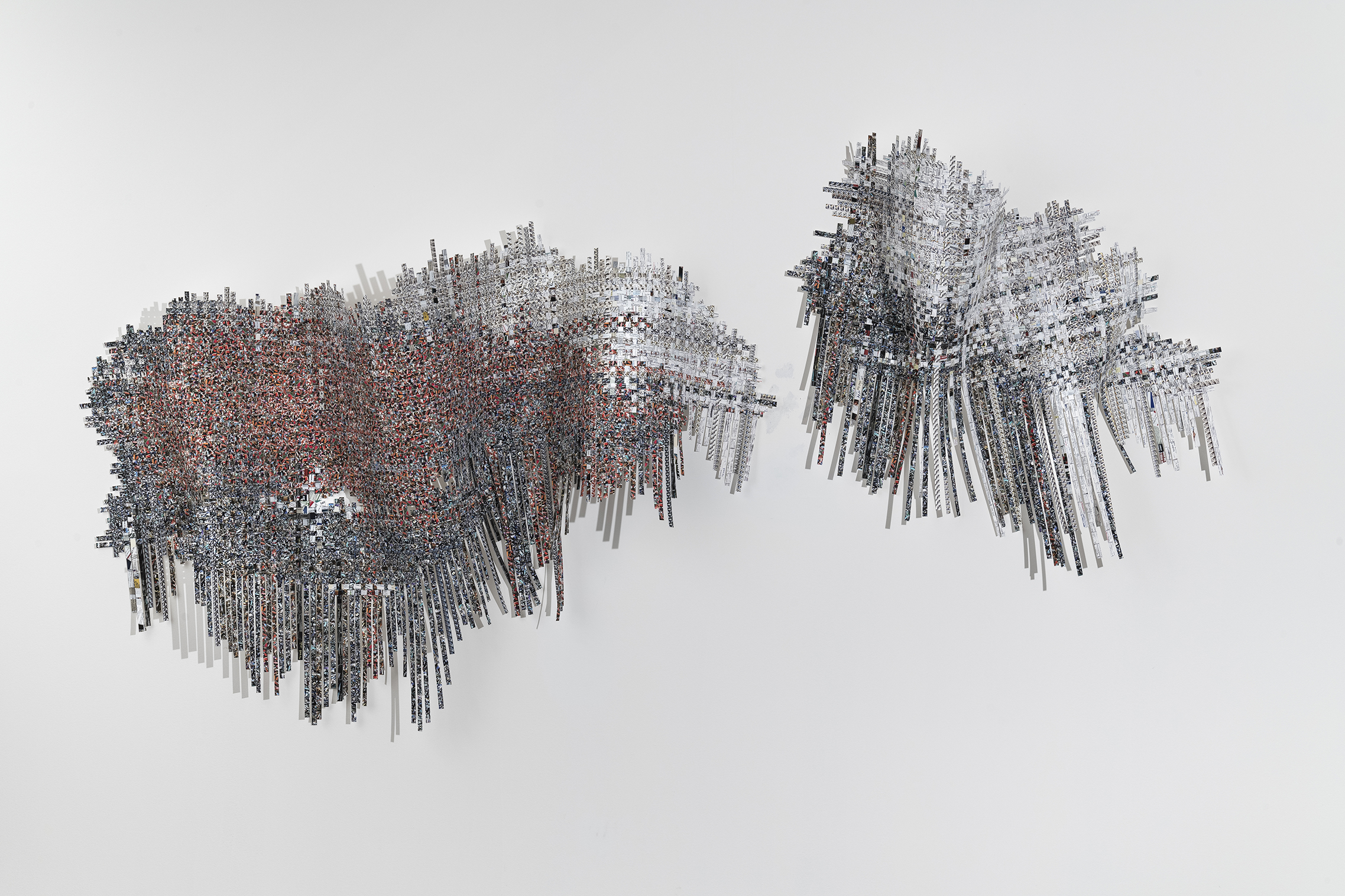 Two abstract sculptural forms composed of densely layered rectangular strips and particles suspended in space against a neutral background. The left form features warm reddish-brown and gray tones with a more compact, cloud-like density, while the right form displays predominantly gray and white tones in a more dispersed, wispy configuration. Both structures create depth through their three-dimensional arrangement of fragmented materials.