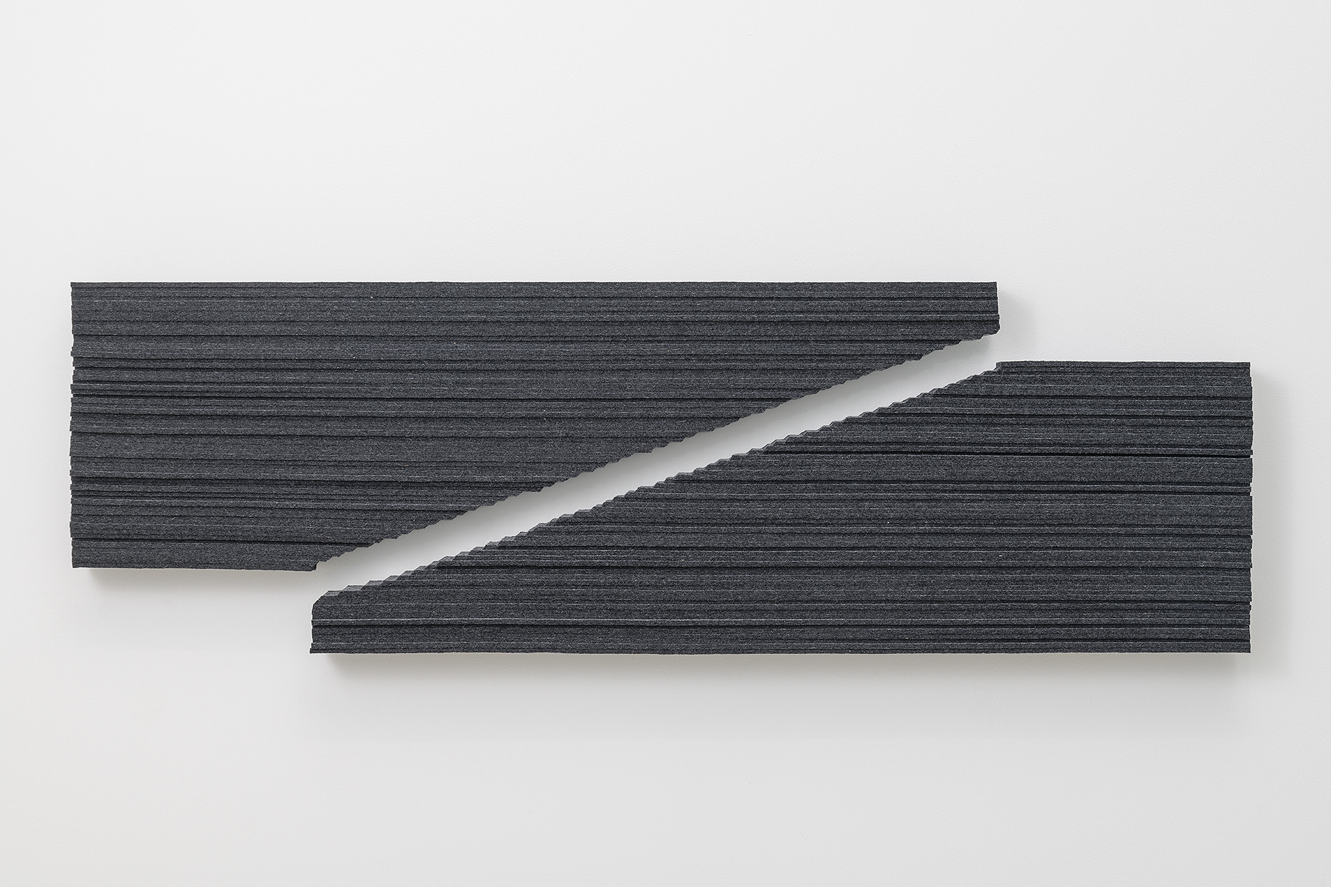 Two dark gray textile strips with horizontal ribbed texture are mounted on a white wall, angled diagonally across one another to create an interlocking geometric composition with a white gap between them.