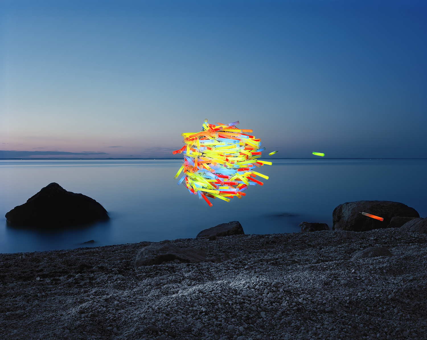 Glowing neon-colored sticks arranged in a chaotic, floating mass above calm twilight waters, photographed from a rocky beach at dusk with silhouetted boulders in the foreground and distant shoreline.
