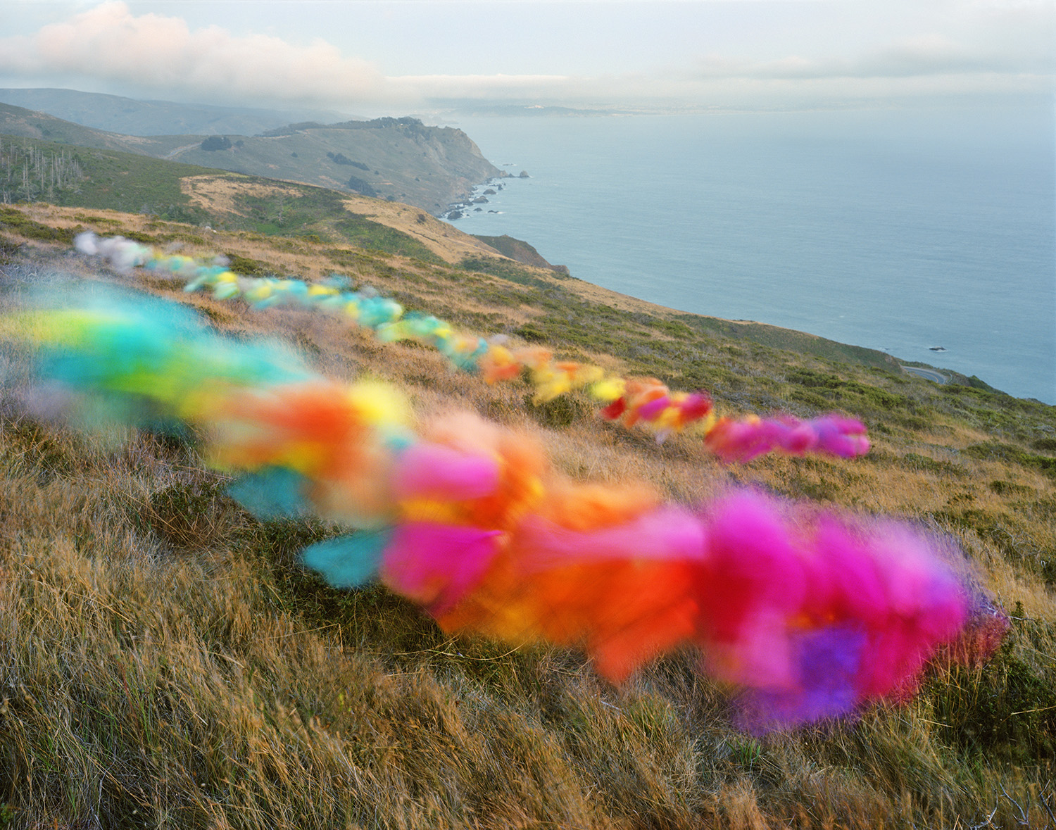 Vibrant rainbow-colored tulle fabric trails across a coastal hillside, creating a striking contrast against dry golden grasses. The composition frames a dramatic seascape beyond, with rocky cliffs, turquoise waters, and a distant coastline under a partly cloudy sky.