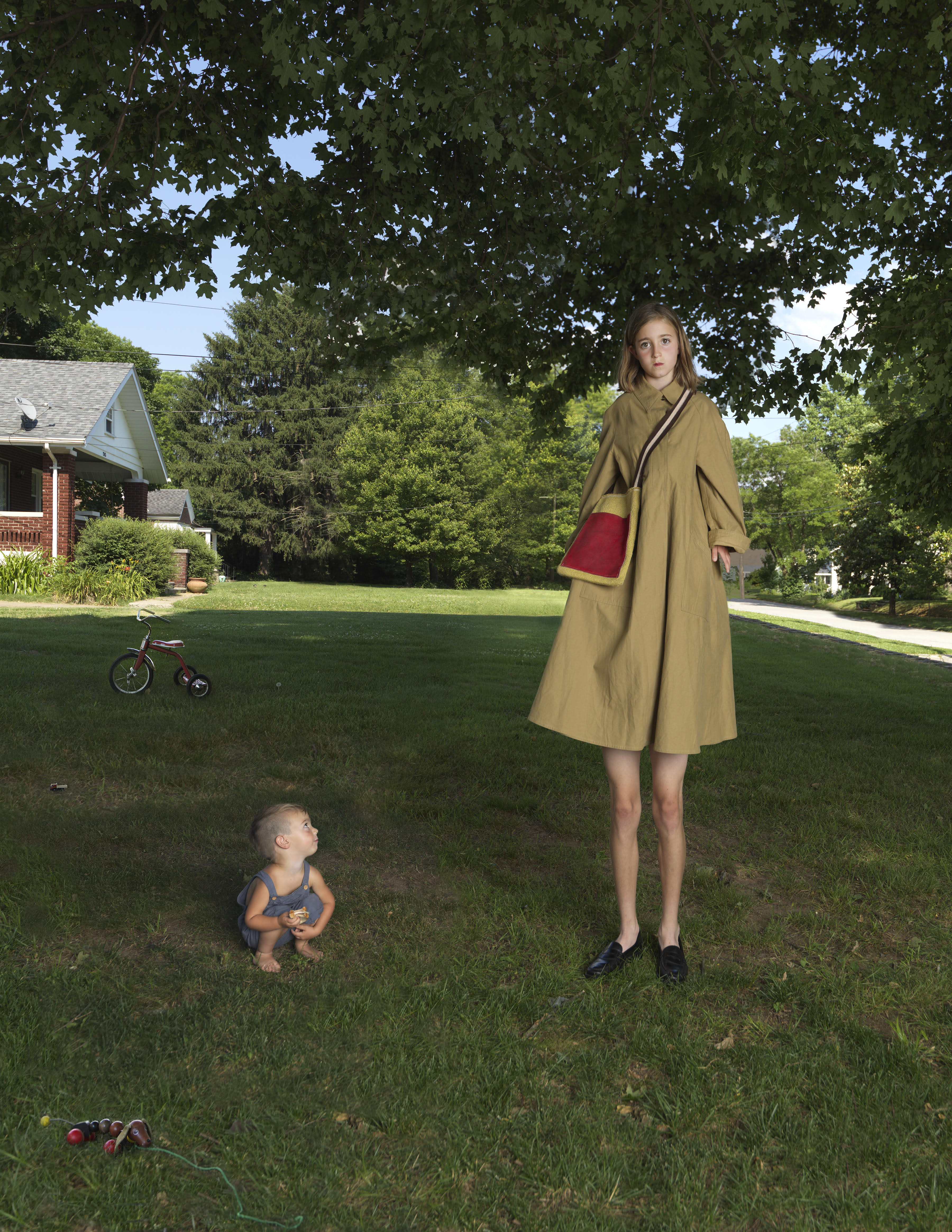 A woman of unnaturally elongated proportions stands in a suburban lawn wearing a tan coat with a burgundy pocket, her impossibly long legs extending to black shoes. A toddler crouches beside her in the grass while a tricycle and flowers sit scattered in the foreground, with a brick house and tree-lined neighborhood visible behind them.