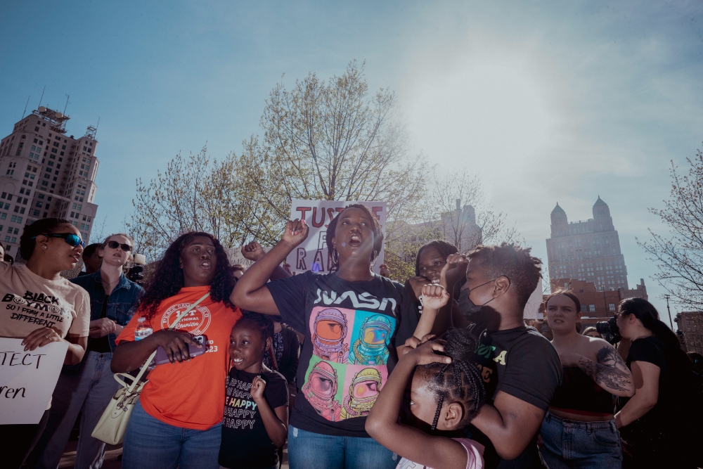 A diverse group of people gather in an urban plaza for a protest, with bare trees and city buildings visible in the background. The crowd includes adults and children, some holding signs, with participants wearing casual clothing in various colors including orange, black, and neutral tones.
