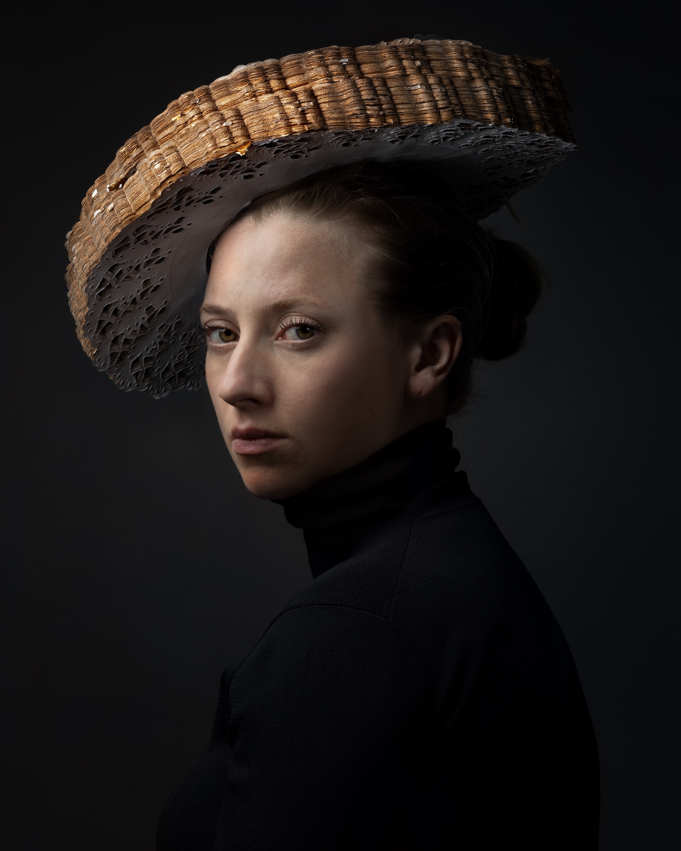 Portrait of a woman in profile against a dark background, wearing a black turtleneck and an elaborate woven hat with a straw-colored crown and dark perforated brim that extends dramatically to the side.