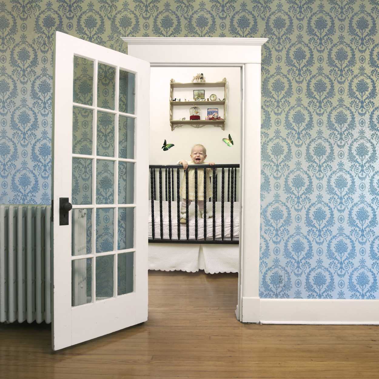 A laughing baby stands in a black crib viewed through an open white glass-paned door, surrounded by damask-patterned wallpaper in shades of blue and green. Decorative shelves and butterfly cutouts are visible on the nursery wall above the crib.