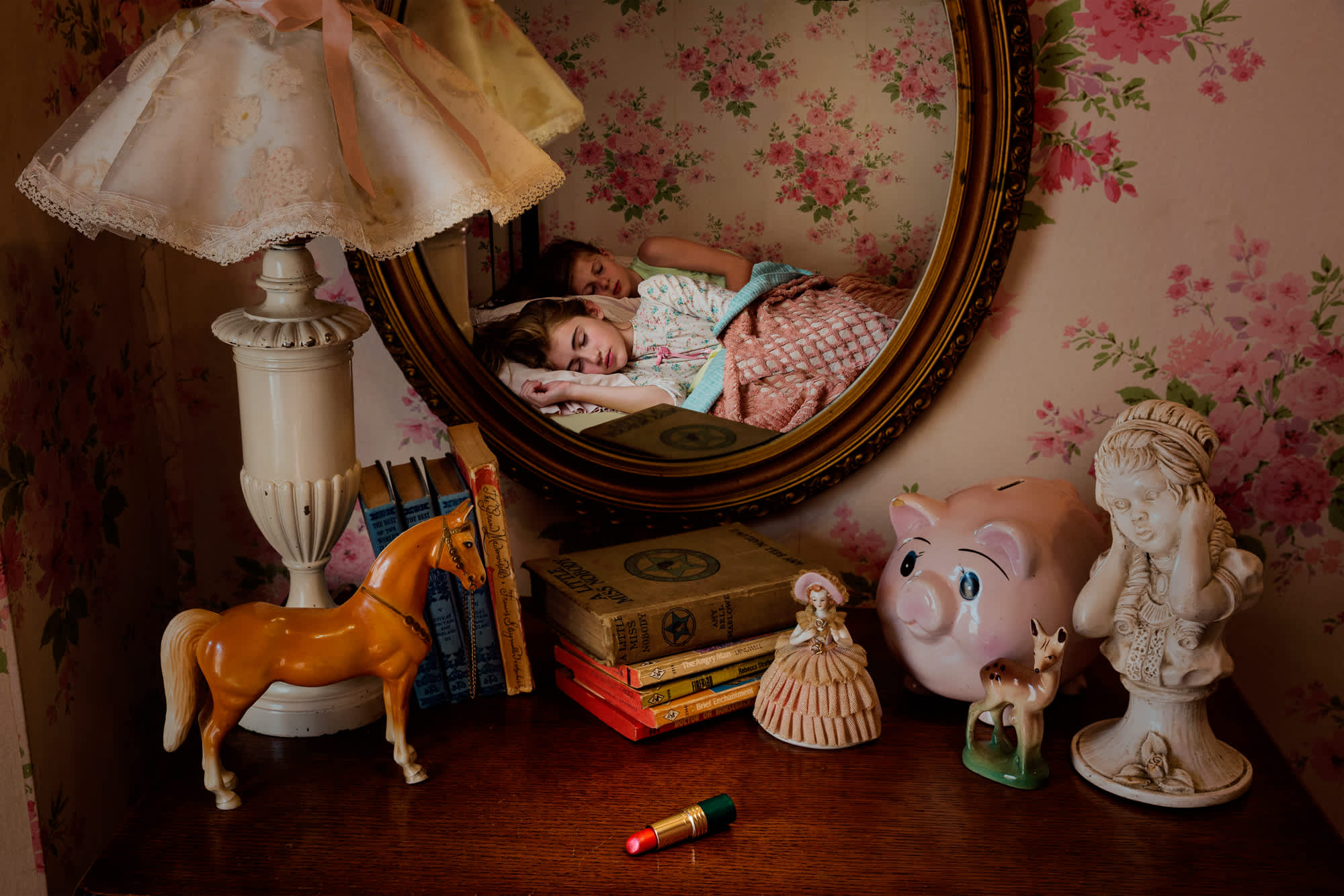 A vintage-styled bedroom scene viewed through an ornate oval mirror, showing two children sleeping in a bed with pink bedding. The foreground displays a wooden dresser arranged with nostalgic objects including a ceramic lamp with lace shade, brown horse figurine, stack of antique books, decorative bust, piggy bank, small figurines, and a red lipstick, against floral wallpaper in muted tones.