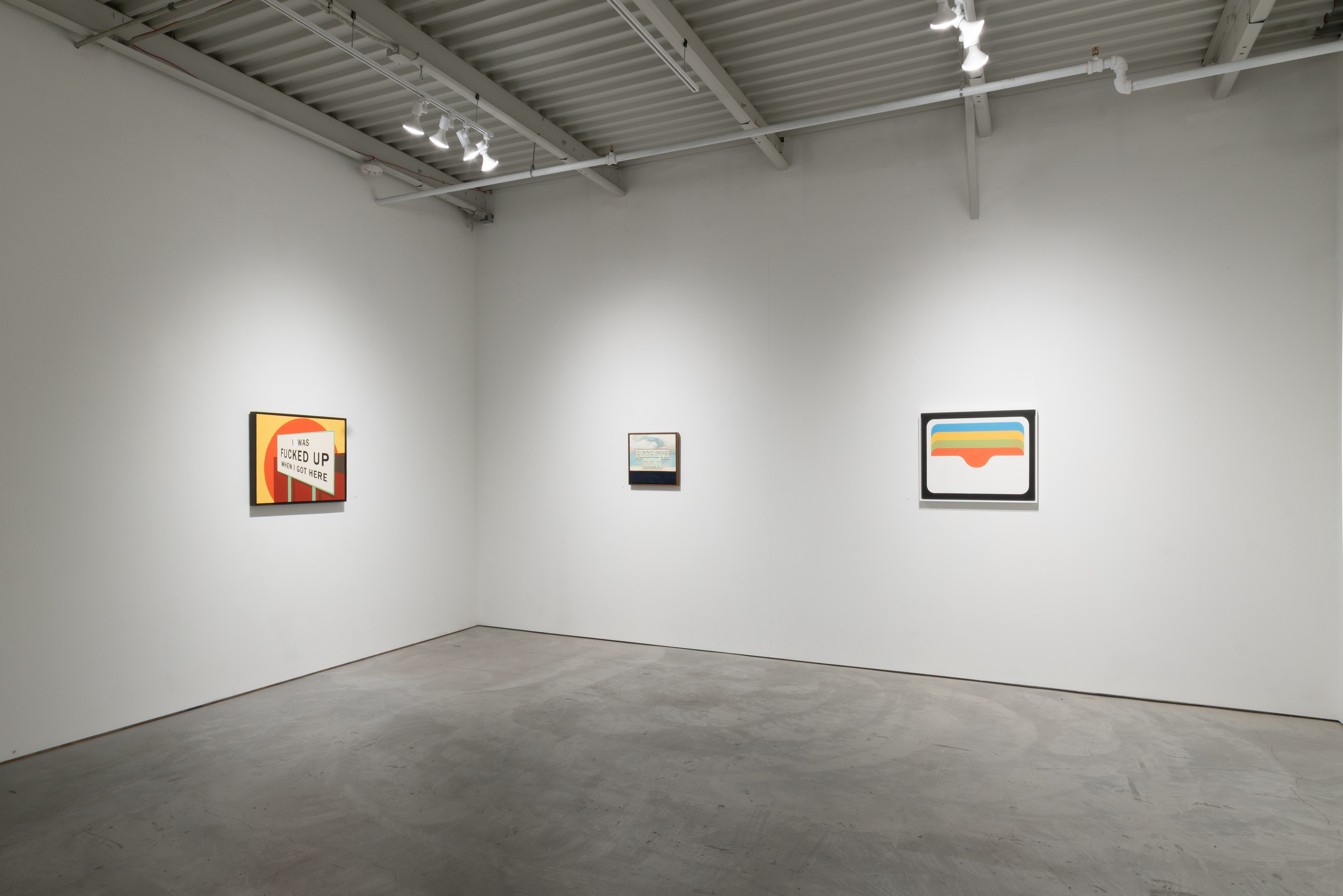 A sparse gallery space with three framed artworks hung on white walls at varying heights. On the left, a piece with bold red and yellow geometric forms and black text; in the center, a smaller work with muted tones; on the right, a composition featuring horizontal stripes in blue, yellow, and orange. Polished concrete floor and industrial ceiling with track lighting.