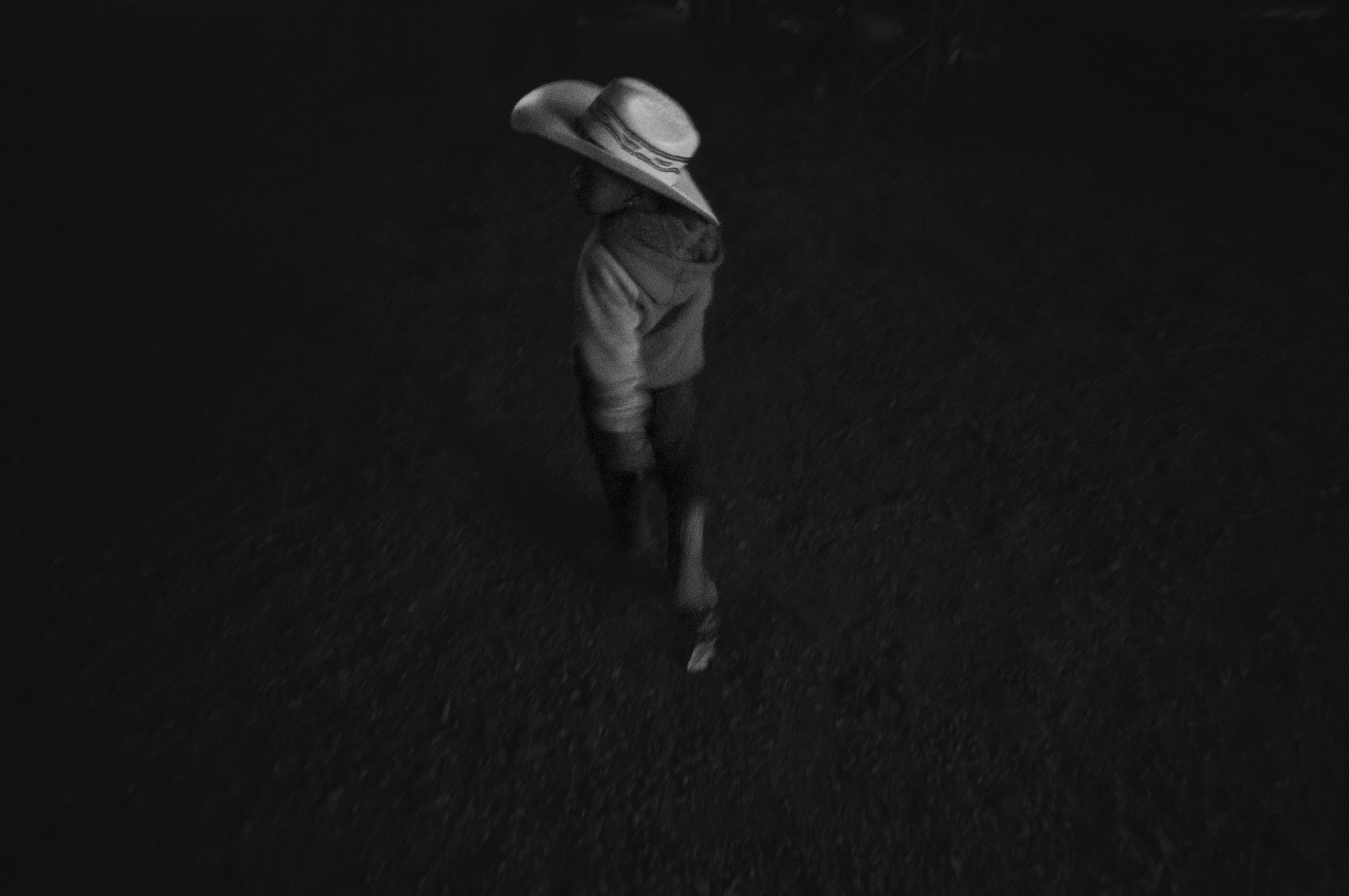 A solitary figure wearing a wide-brimmed hat and wrapped in fabric stands against a dark, textured ground in this high-contrast black and white photograph. The composition emphasizes isolation and stillness, with the figure emerging from shadow in a minimalist, enigmatic composition.