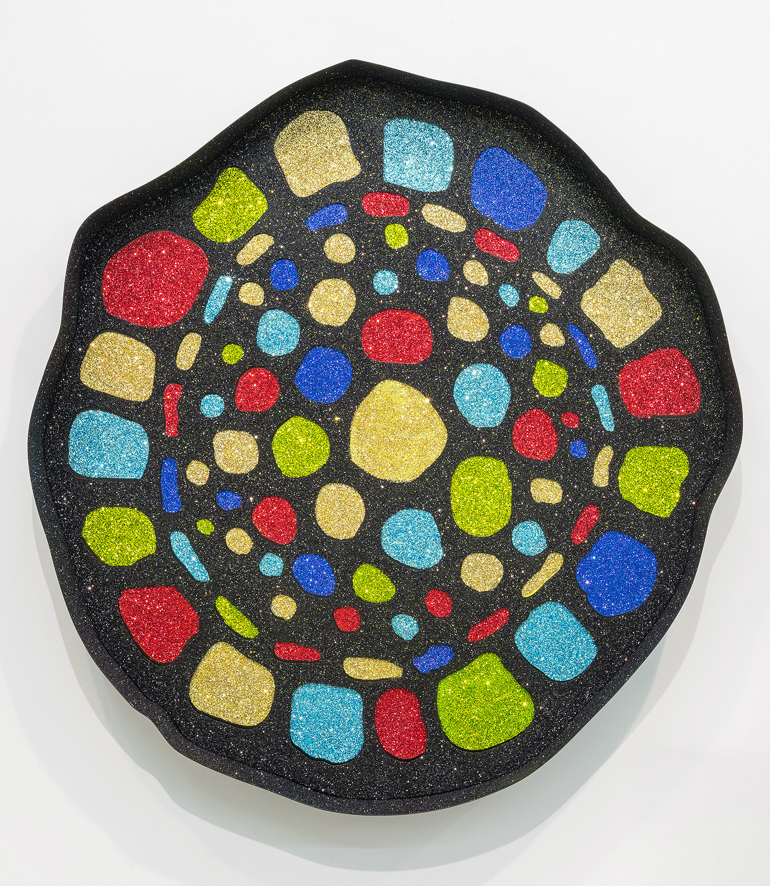An organic, kidney-shaped black panel densely covered with colorful glittering shapes in red, yellow, blue, cyan, and lime green. The varied forms—ranging from small circles and rectangles to larger rounded and squared shapes—are scattered across the surface with a shimmering, jewel-like finish that creates an energetic, mosaic-like composition.