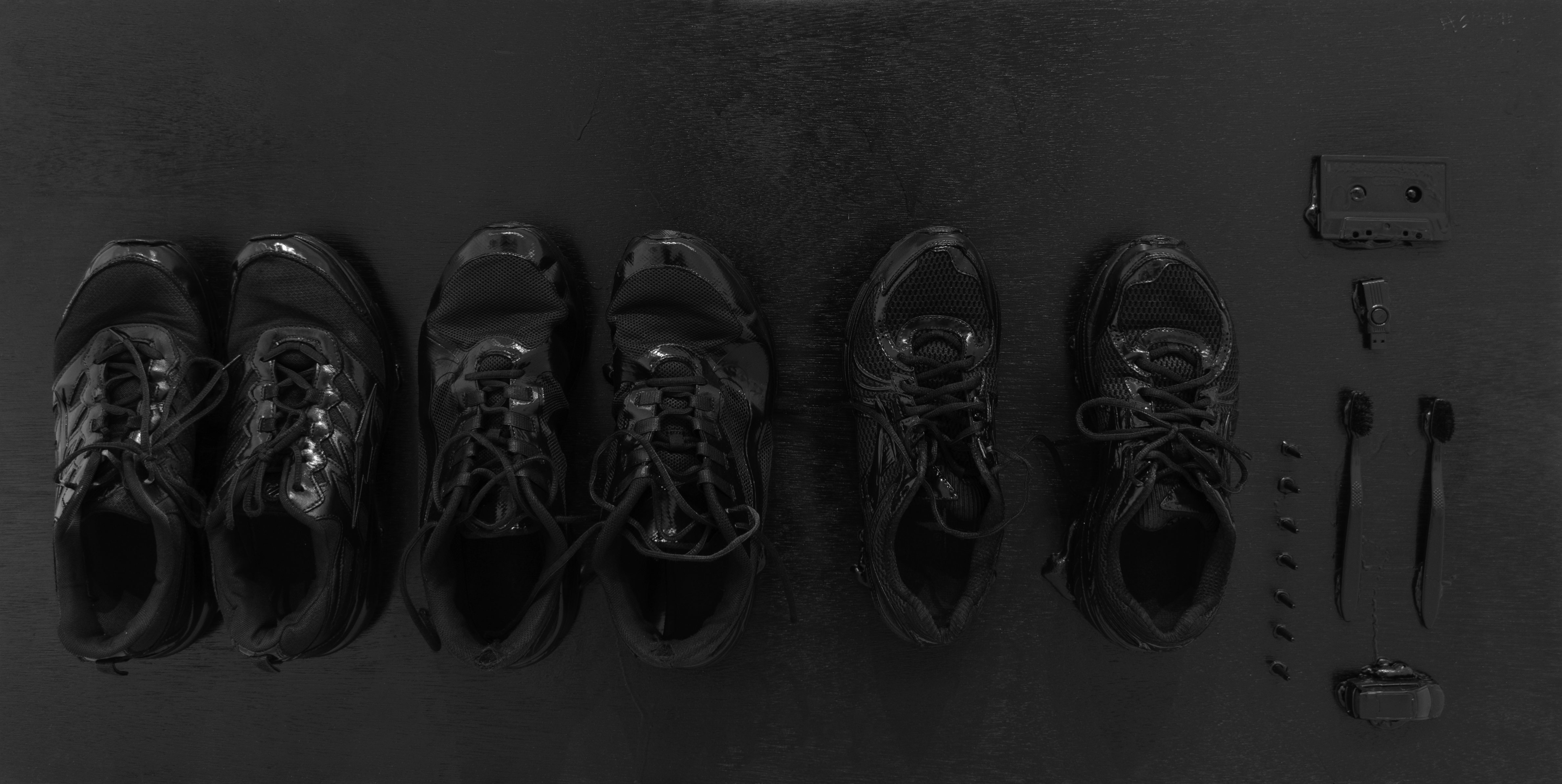 Six black athletic shoes arranged in pairs on a dark wooden surface, accompanied by scattered objects including bullets, toothbrushes, a cassette tape, and a USB drive arranged to the right. The monochromatic composition uses found objects and everyday items to create a stark, documentary-style arrangement against a textured dark background.