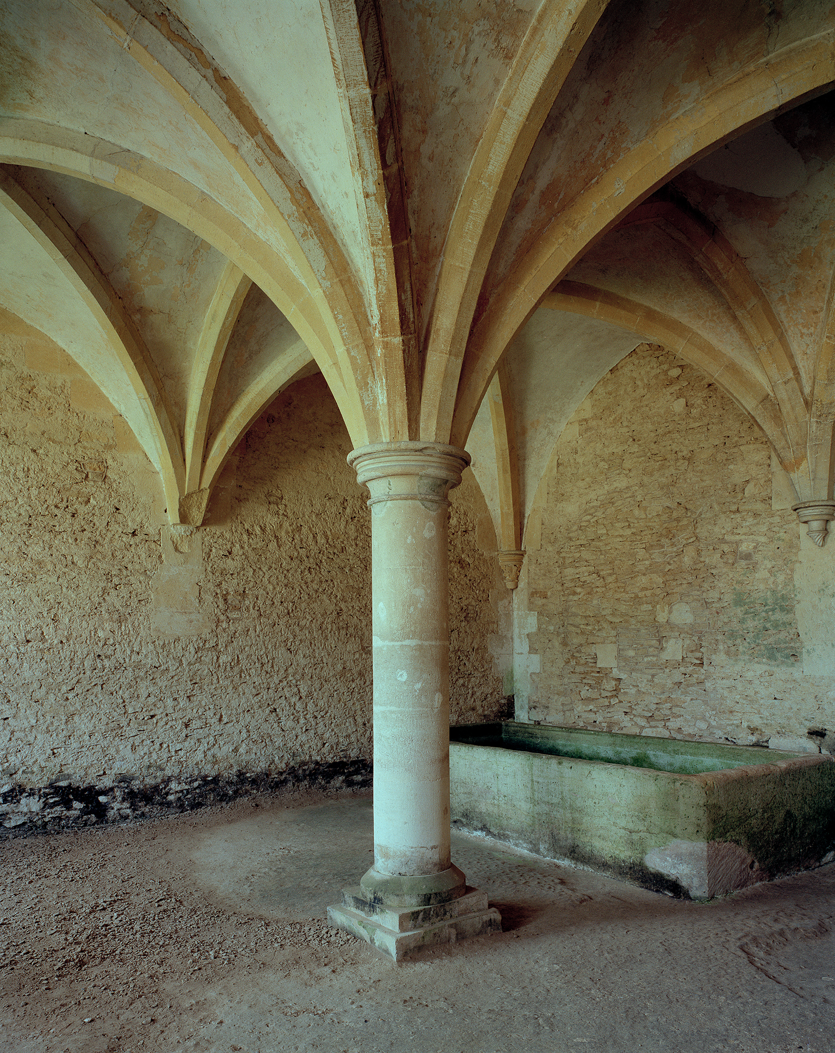 Vaulted stone corridor with elegant ribbed arches supported by a central cylindrical column. Weathered cream and tan masonry walls show age and patina, with a green-tinted water channel running along the right side of the dusty earthen floor.