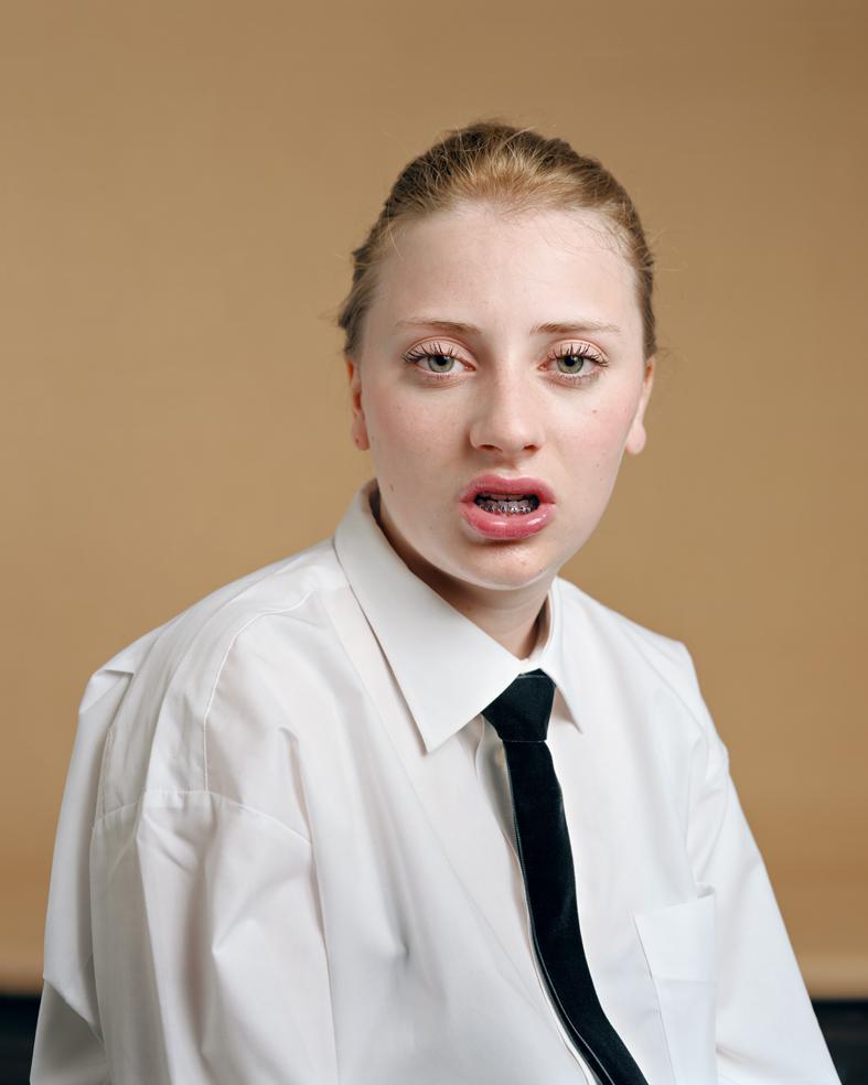 Portrait of a young person with reddish-blonde hair pulled back, wearing a white collared shirt and black tie against a warm tan background. The subject gazes directly at the viewer with a slightly open mouth expression, rendered in vivid color with sharp photographic clarity.