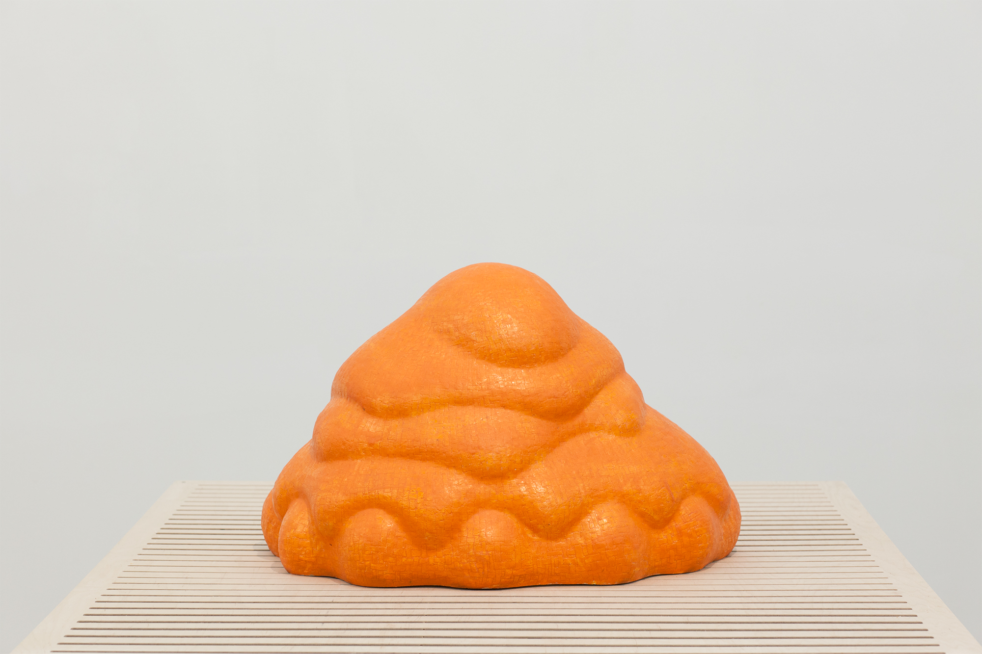 A bright orange glazed ceramic sculpture with an organic, rounded form sits on a striped wooden platform. The bulbous shape features layered, undulating surfaces that suggest both natural and abstract qualities against a neutral gray background.