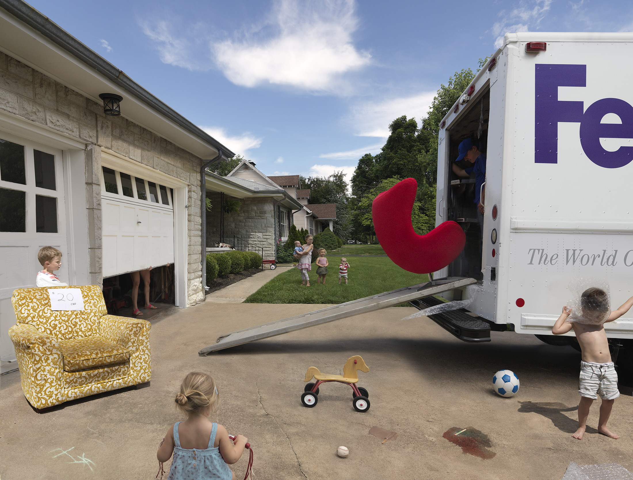 A suburban driveway scene depicts children at play around a yellow patterned armchair and delivery truck with a large red sculptural form protruding from its open cargo door. Additional children play in the grassy yard behind, while a residential home with white garage and stone facade frames the left side of the composition.