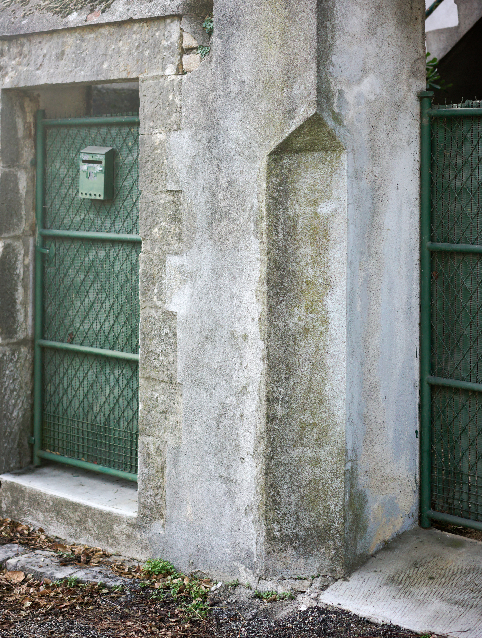 Weathered concrete pillars and walls marked by moss and lichen staining frame a narrow passageway lined with green metal gates and a mailbox, capturing the texture and decay of an industrial or utilitarian structure.