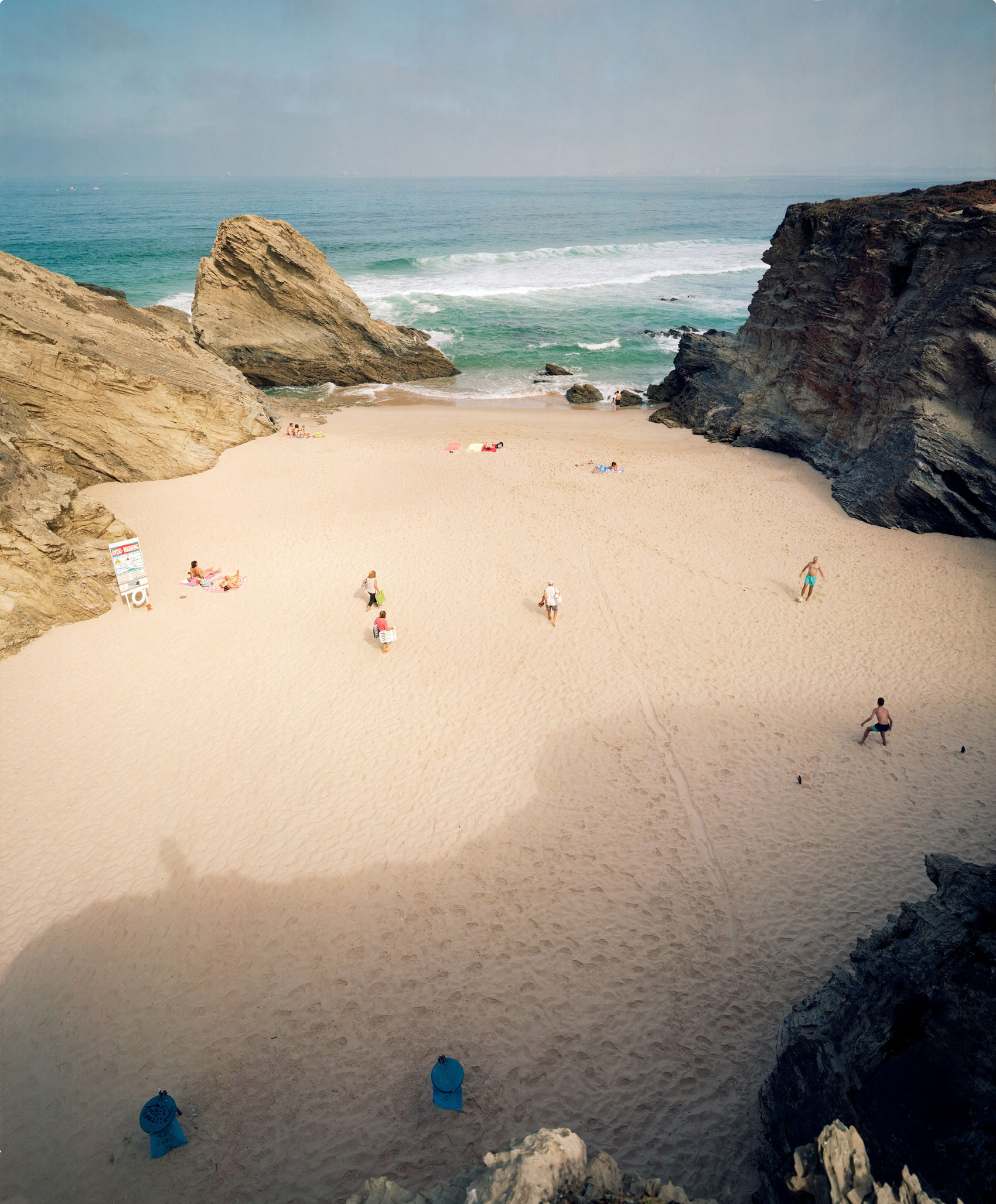 Aerial view of a secluded sandy beach nestled between towering dark rock formations, with turquoise waters and white-capped waves beyond. Scattered beachgoers and blue umbrellas dot the pale sand, creating small focal points across the expansive composition.