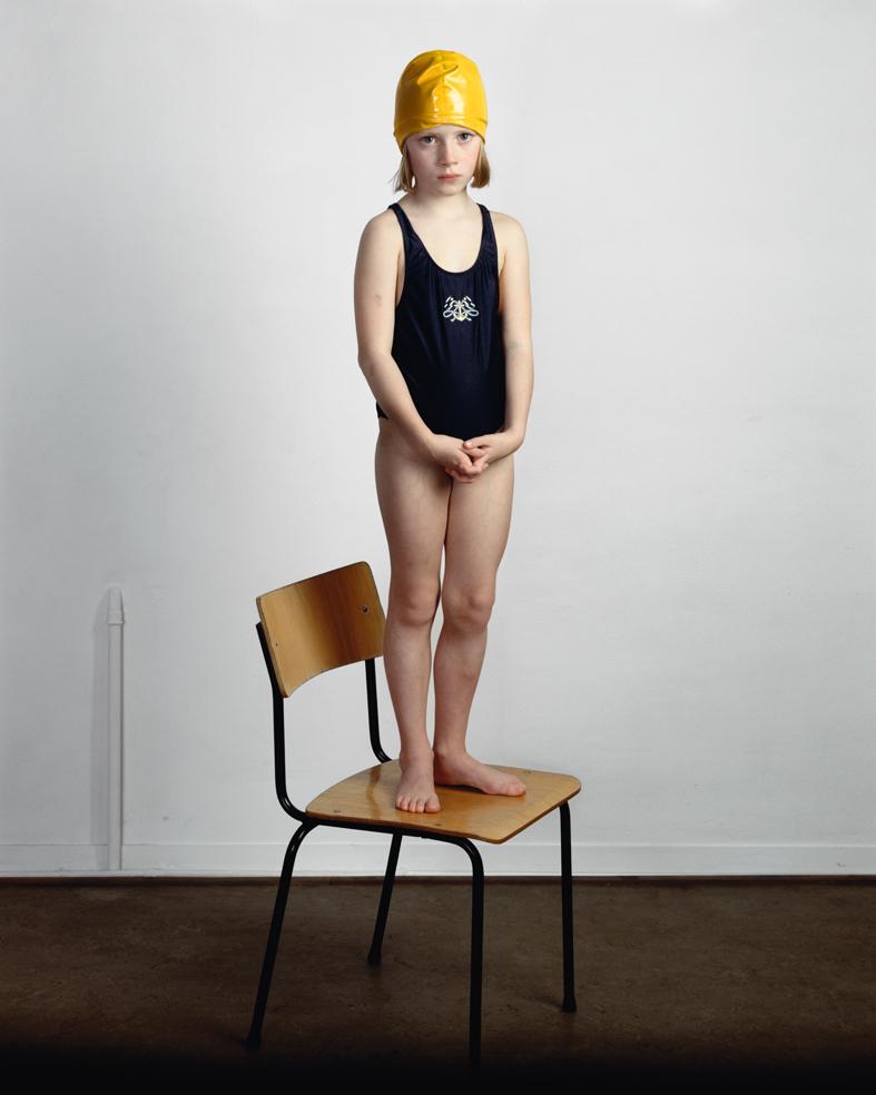 A young child in a black swimsuit and bright yellow swimming cap stands barefoot on a wooden chair with black metal legs, positioned against a white wall. The child's hands are clasped in front, gazing directly at the camera in a contemplative pose.