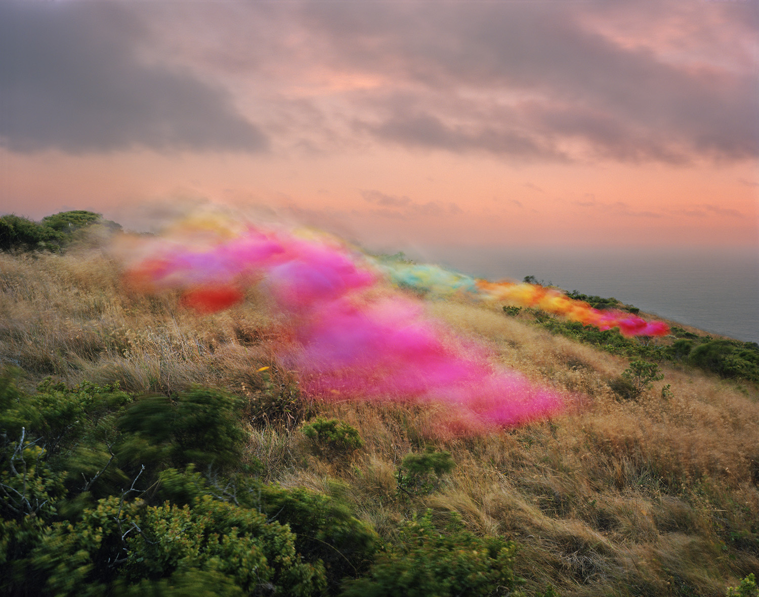 Hillside landscape with dry golden grasses and green shrubs beneath a soft pink and gray sky, overlaid with billowing clouds of magenta, yellow, and orange pigment that drift across the terrain like colored smoke.