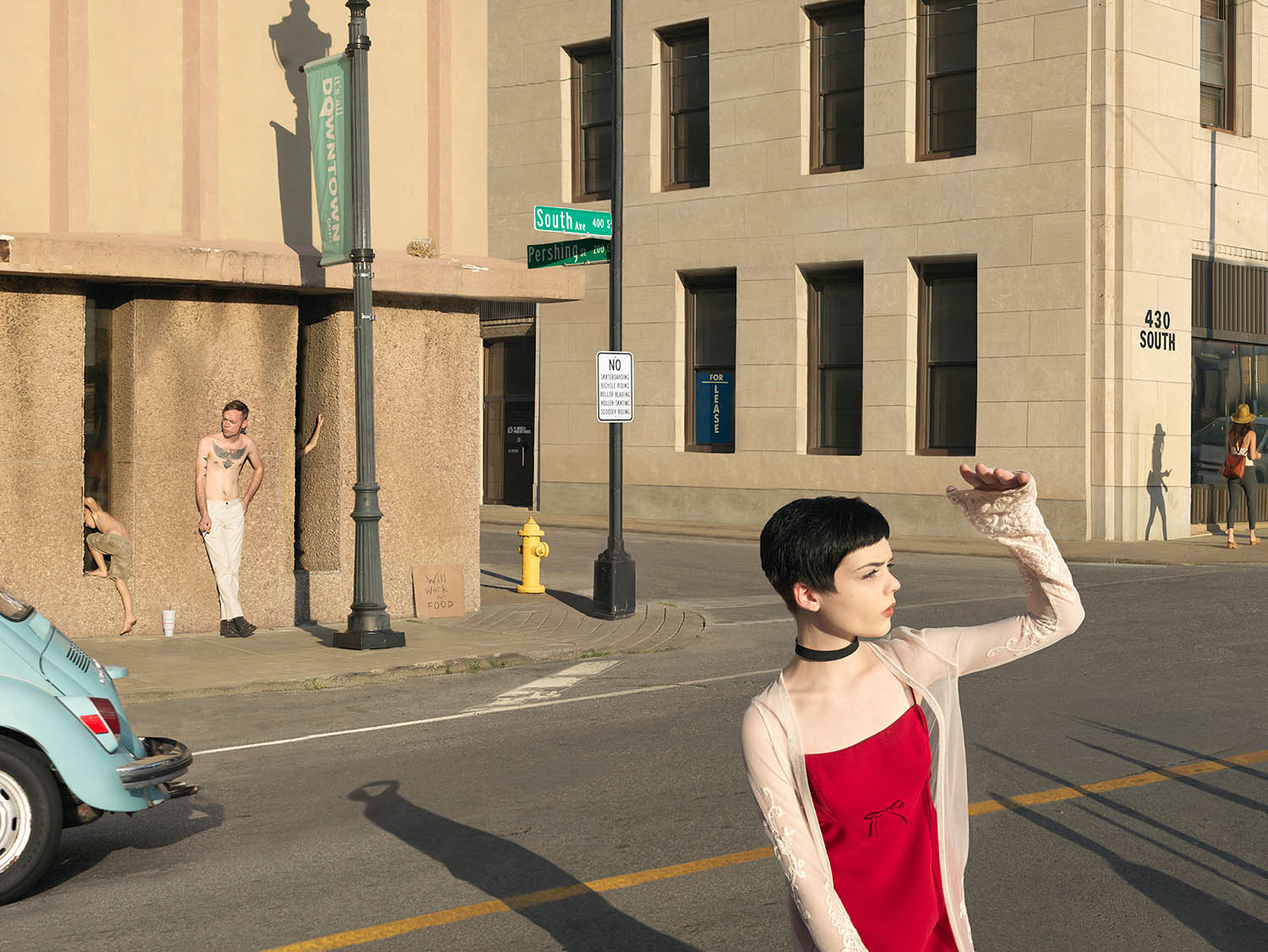 A woman in a red dress and beige cardigan stands in the foreground of an urban street intersection, shading her eyes while looking upward. Behind her, figures in minimal clothing pose against a concrete structure near a street sign marking South Avenue and Pershing Street, while a vintage turquoise car and building facades frame the sunny, shadow-filled scene.